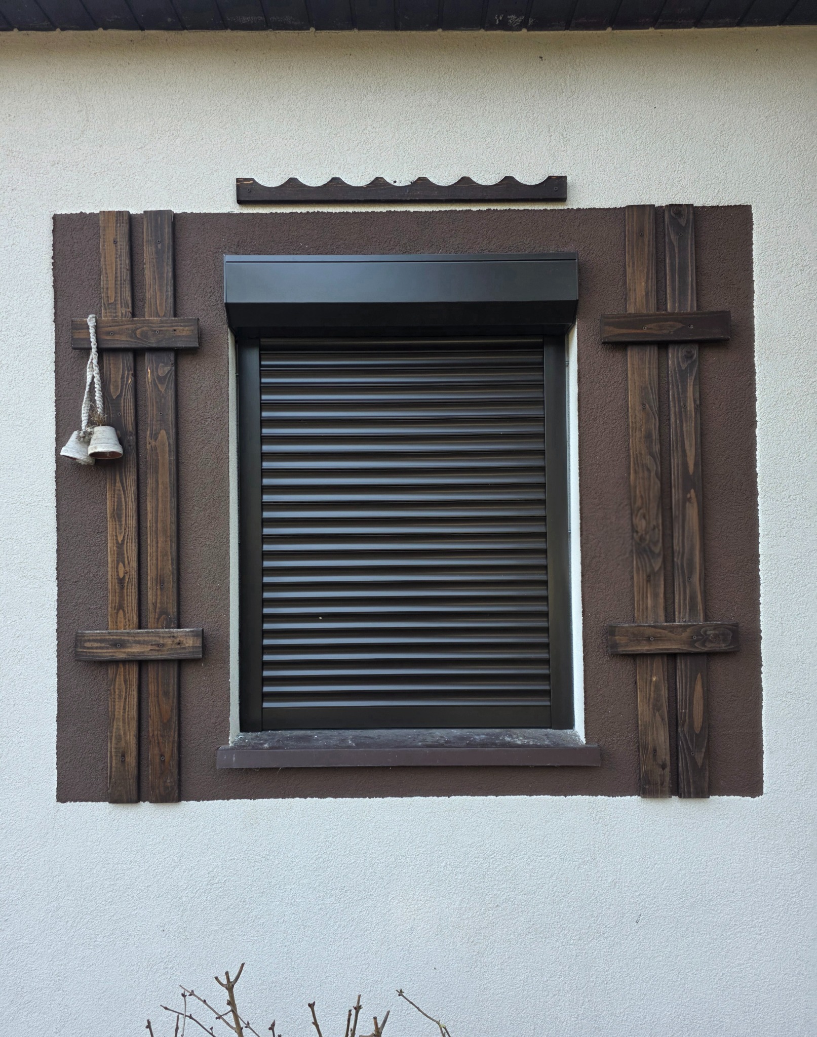 Okno z brązową roletą zewnętrzną, ozdobione drewnianymi elementami i dzwonkami. Elewacja budynku w kolorze białym i brązowym.