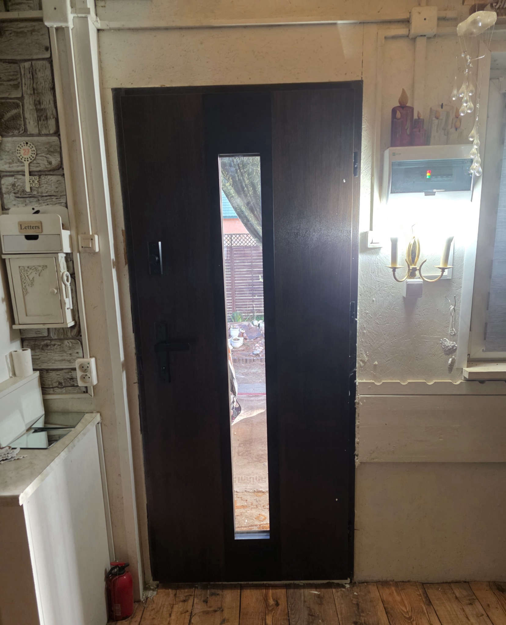 Ciemne drzwi zewnętrzne z wąskim przeszkleniem, widok na ogród. Po lewej skrzynka na listy, po prawej lampa ścienna. Drewniana podłoga, jasne ściany.