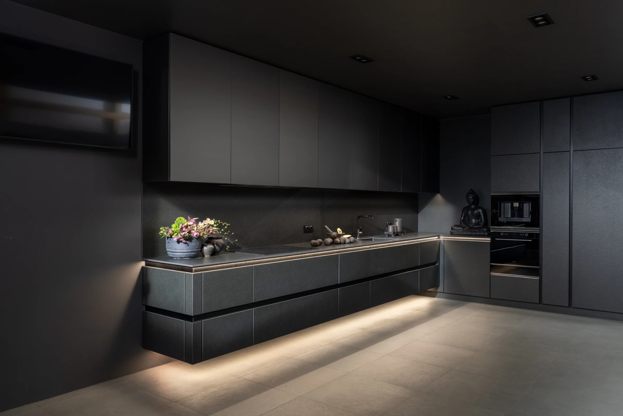 Nowoczesna, minimalistyczna kuchnia w odcieniach szarości z podświetleniem LED pod szafkami. Na blacie dekoracje, w rogu statuetka Buddy. Telewizor na ścianie.