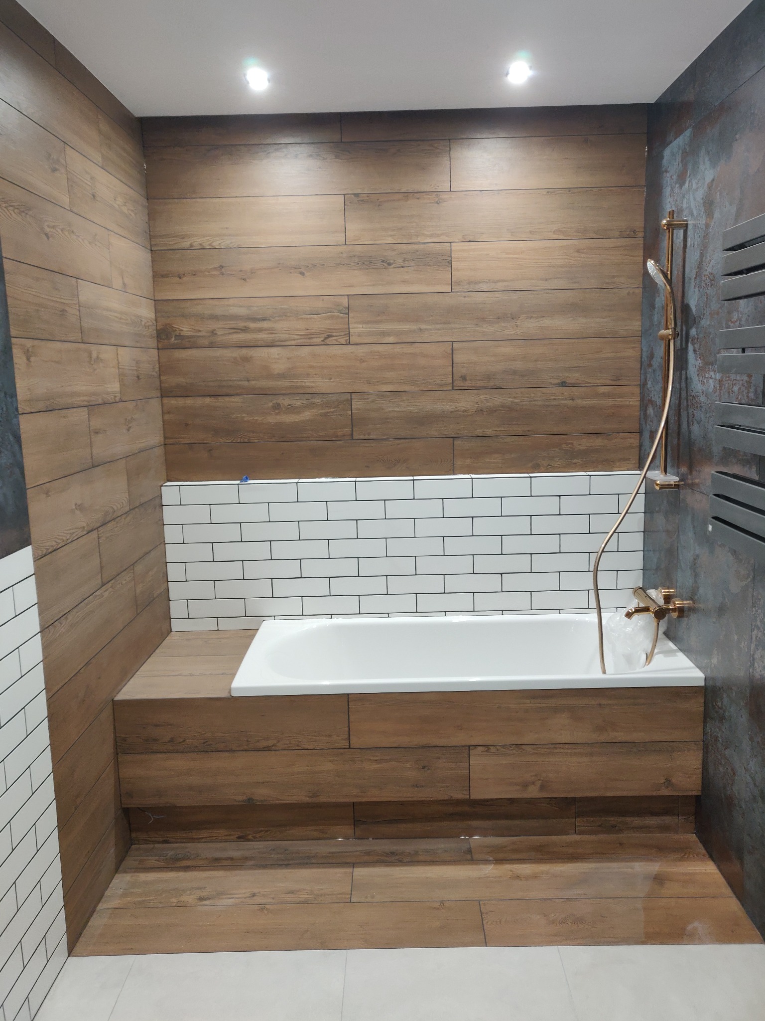 Nowoczesna łazienka z wanną obudowaną drewnopodobnymi płytkami, białe płytki typu metro nad wanną, złota armatura i szara ściana z efektem rdzy.