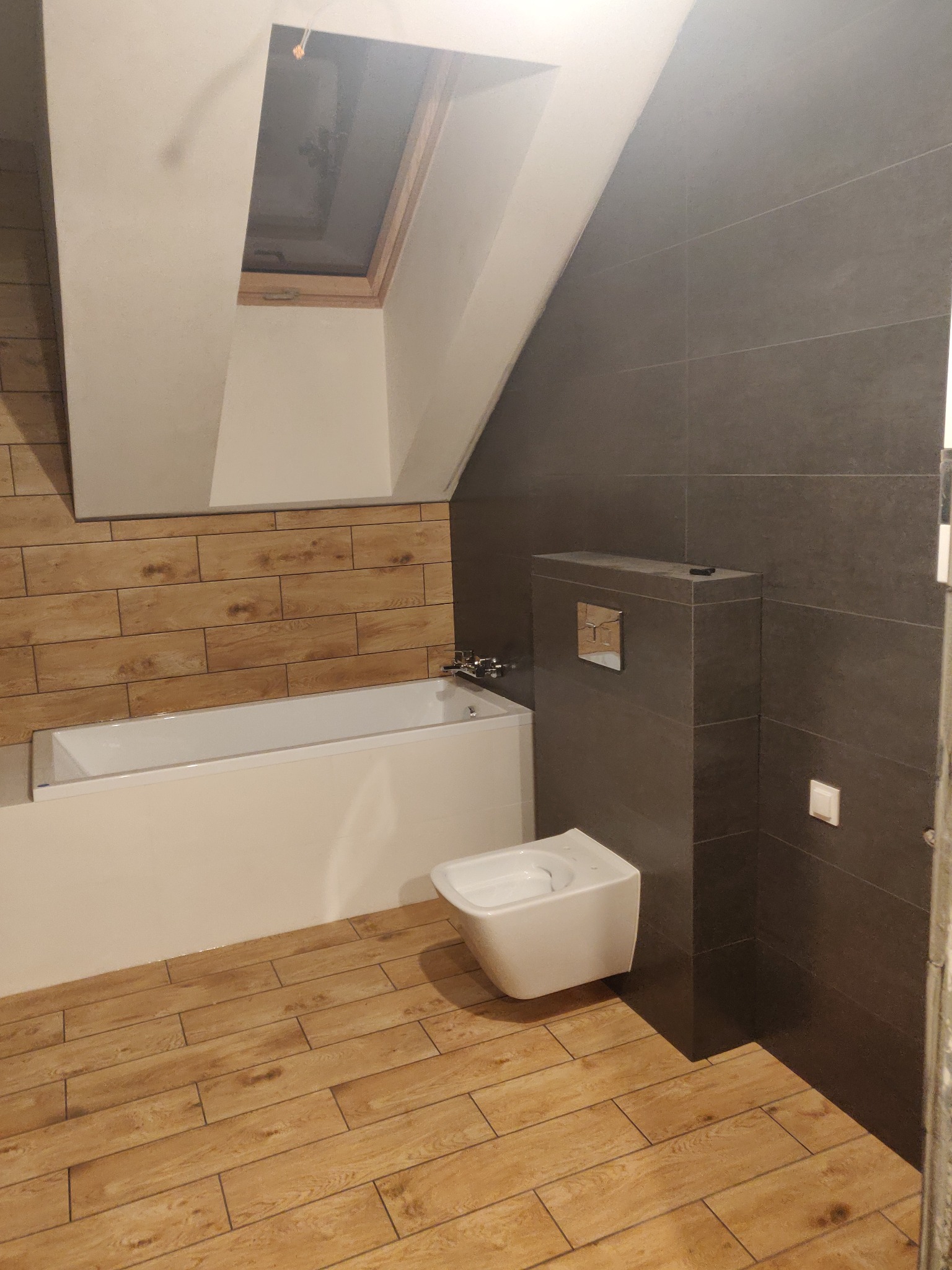 Wykończona łazienka na poddaszu z drewnopodobną podłogą, białą wanną i wiszącym bidetem na tle grafitowych płytek. Okno dachowe doświetla wnętrze.