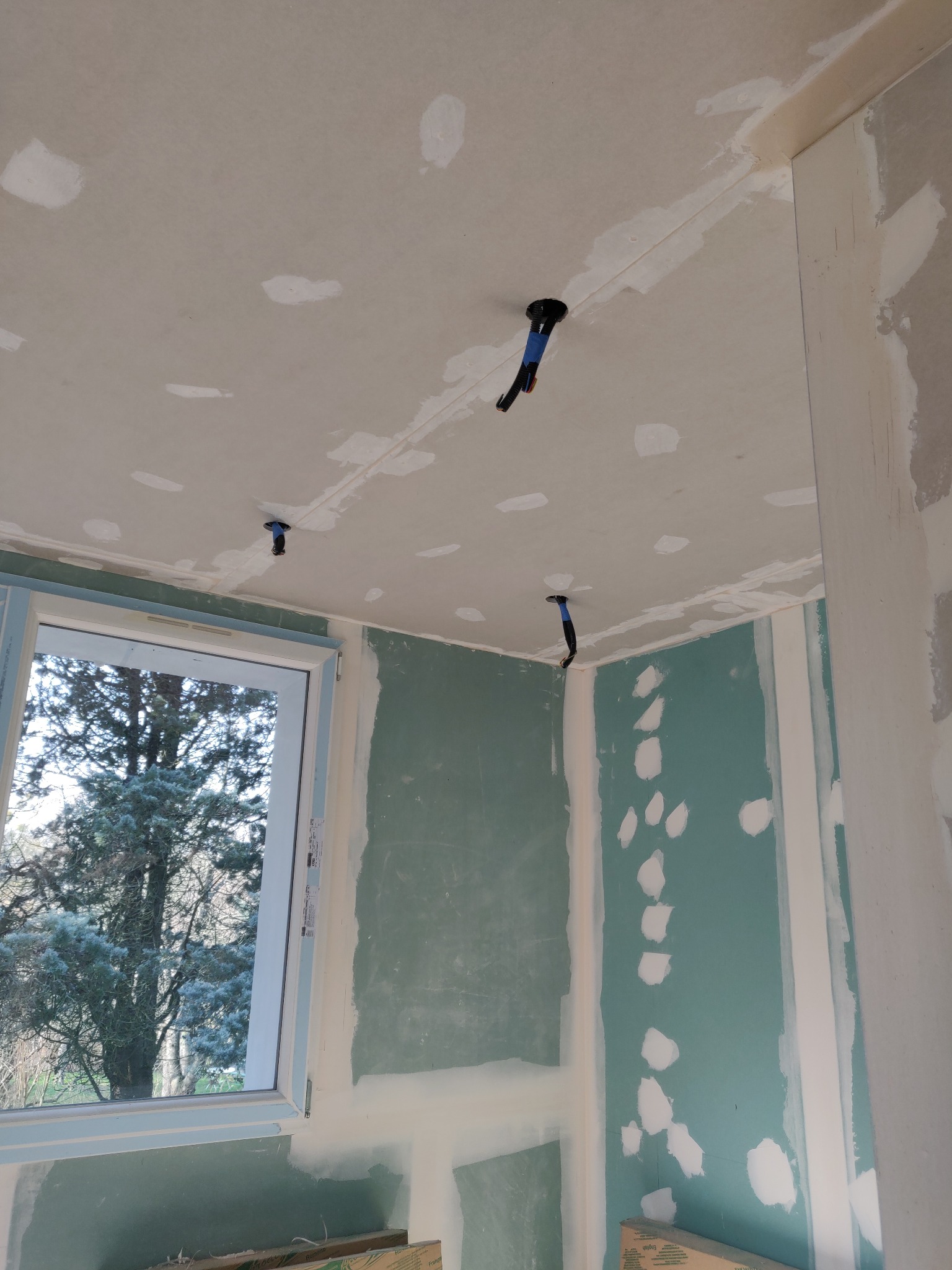 Pokój w trakcie prac wykończeniowych: ściany z płyt gipsowo-kartonowych, widoczne łączenia i otwory po instalacji elektrycznej, okno z widokiem na drzewo.