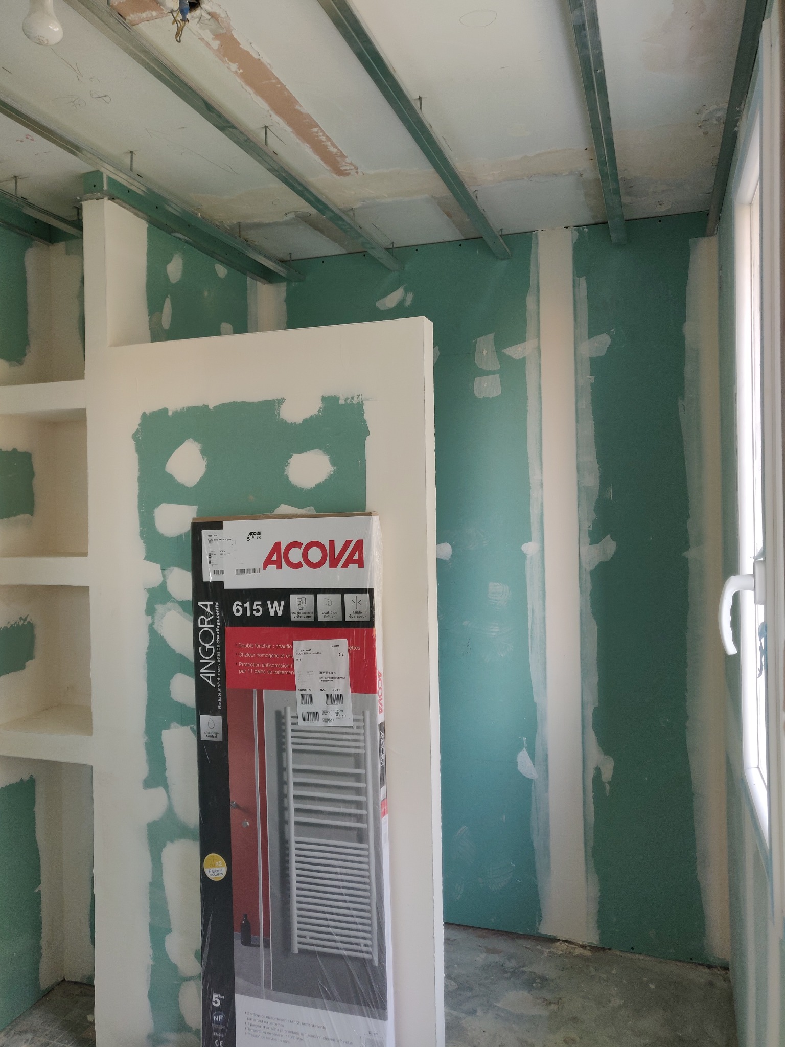 Wnętrze w trakcie prac wykończeniowych: ściany z płyt karton-gips, widoczne profile sufitowe i opakowanie grzejnika łazienkowego Acova Angora 615W.