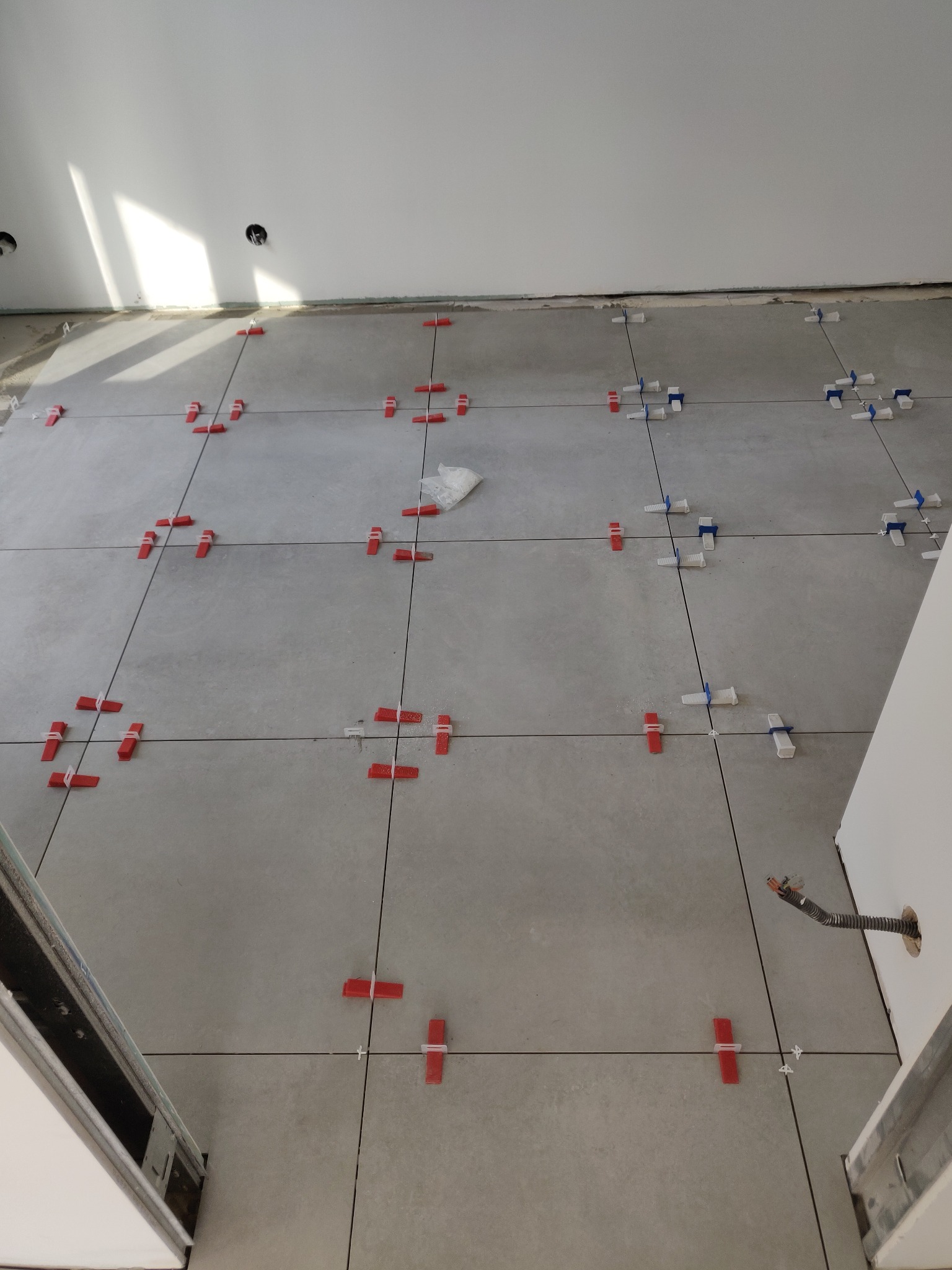 Szare płytki podłogowe, świeżo ułożone, z czerwonymi i białymi klipsami poziomującymi. Widoczny fragment ściany z puszkami elektrycznymi i przewód w peszlu.