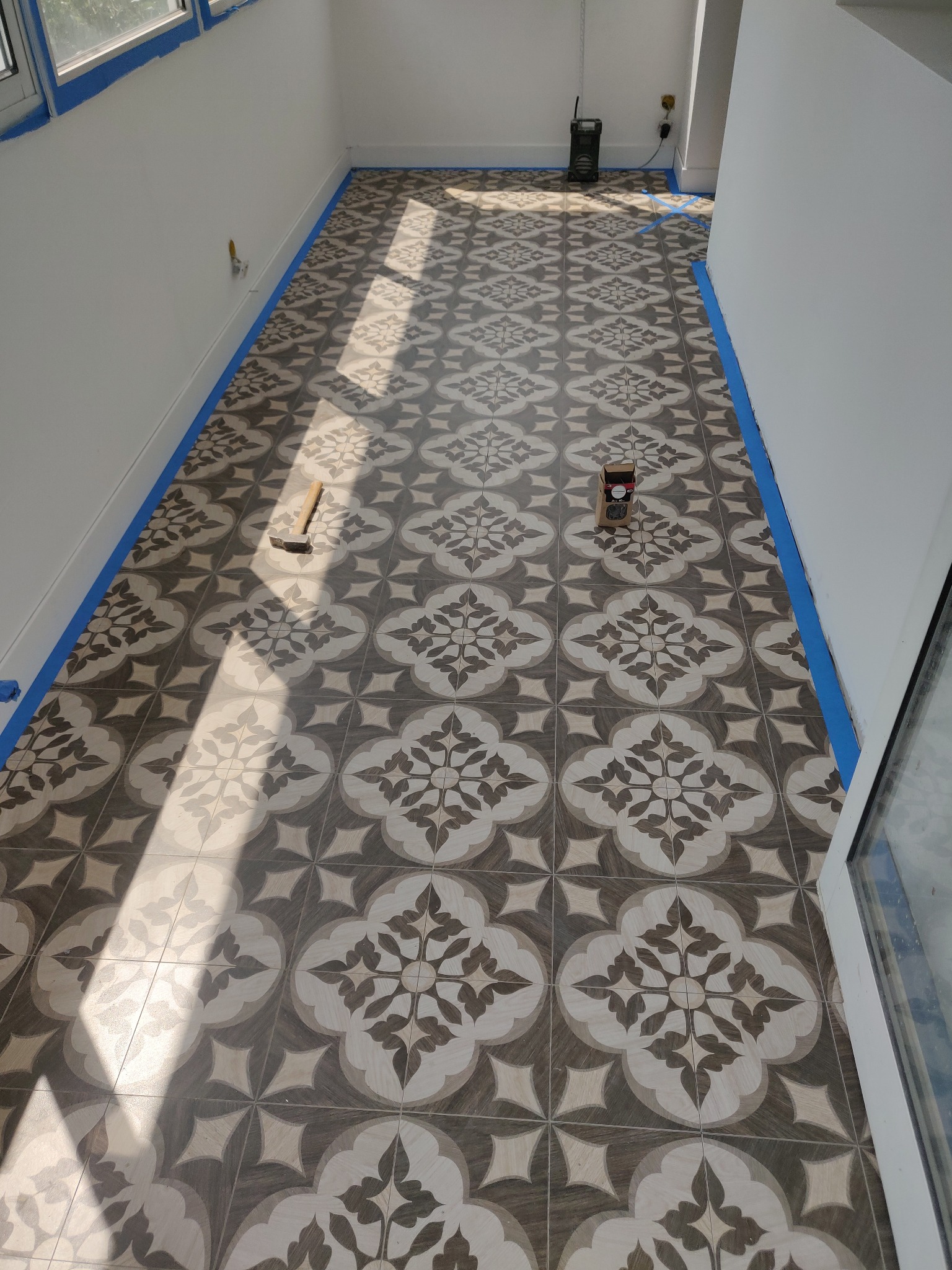 Wnętrze z nową, wzorzystą podłogą w odcieniach szarości i beżu, zabezpieczone taśmą malarską przy ścianach. Widoczny młotek i pudełko po płytkach. Promienie słoneczne podkreślają wzór.