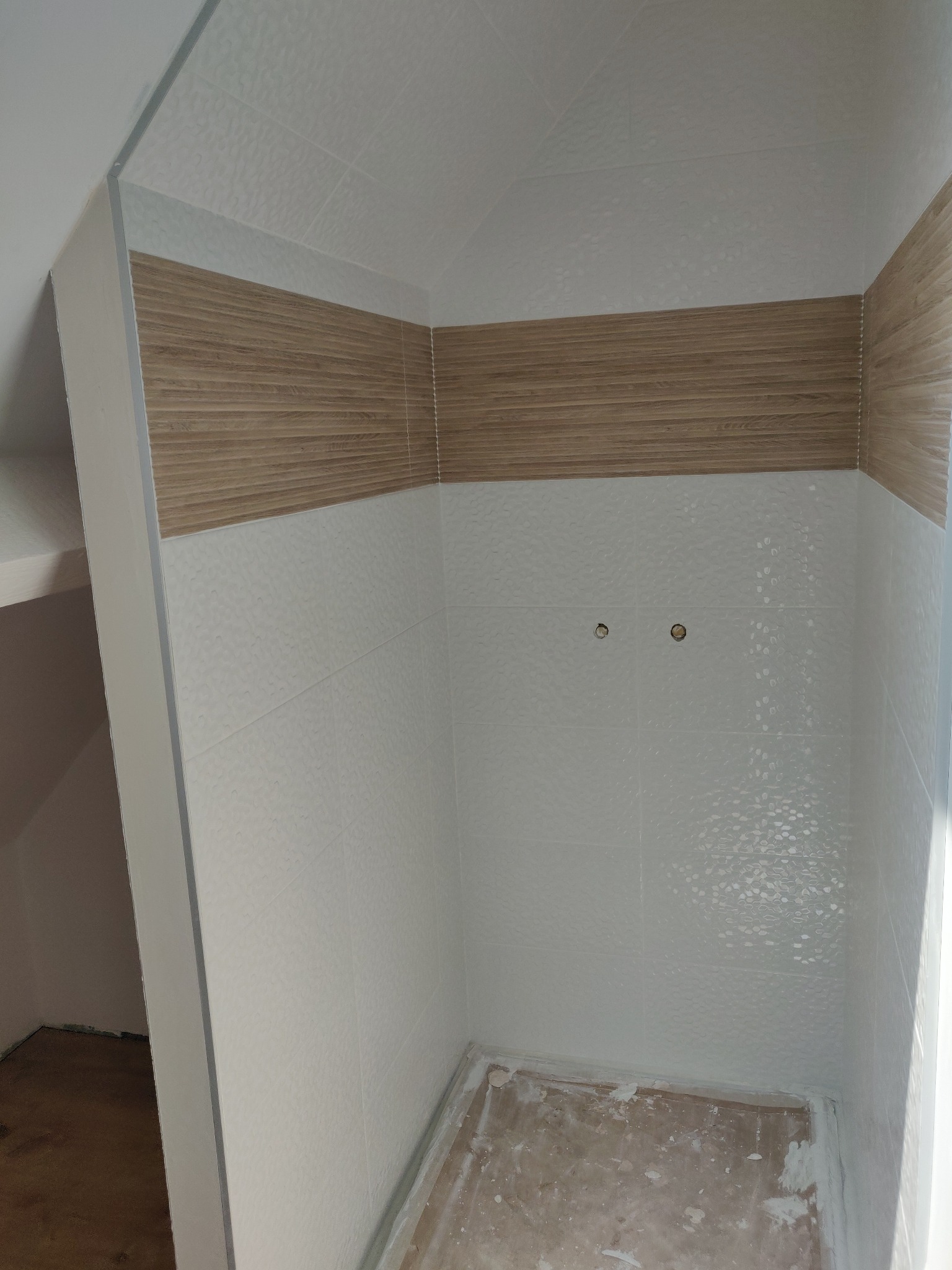 Wykończona wnęka prysznicowa z białymi płytkami o delikatnej fakturze i poziomym pasem dekoracyjnym imitującym drewno. Widoczne przygotowane otwory na armaturę.