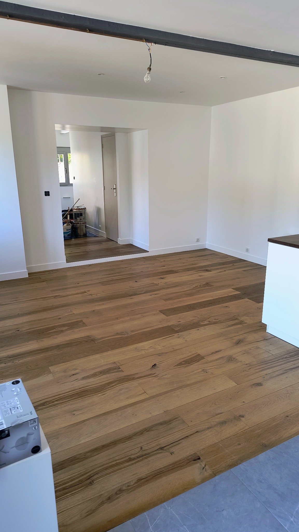 Jasne, odnowione wnętrze z drewnianą podłogą i świeżo pomalowanymi ścianami. Widoczne przejście do kolejnego pomieszczenia i fragment sprzętu remontowego.