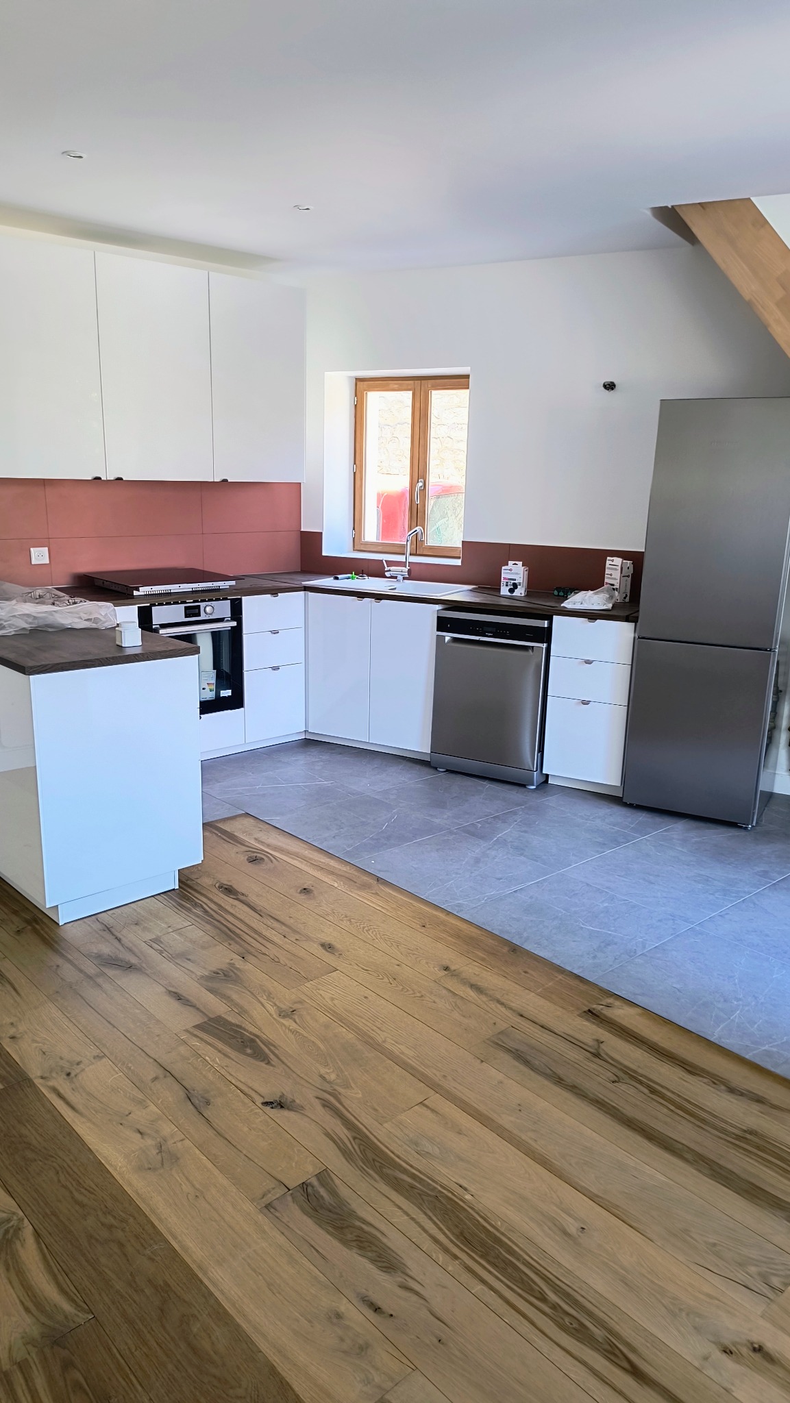 Jasna kuchnia z białymi szafkami, brązowym pasem nad blatem i drewnianą podłogą. Widoczny sprzęt AGD: lodówka, zmywarka, piekarnik. Okno z drewnianą ramą.