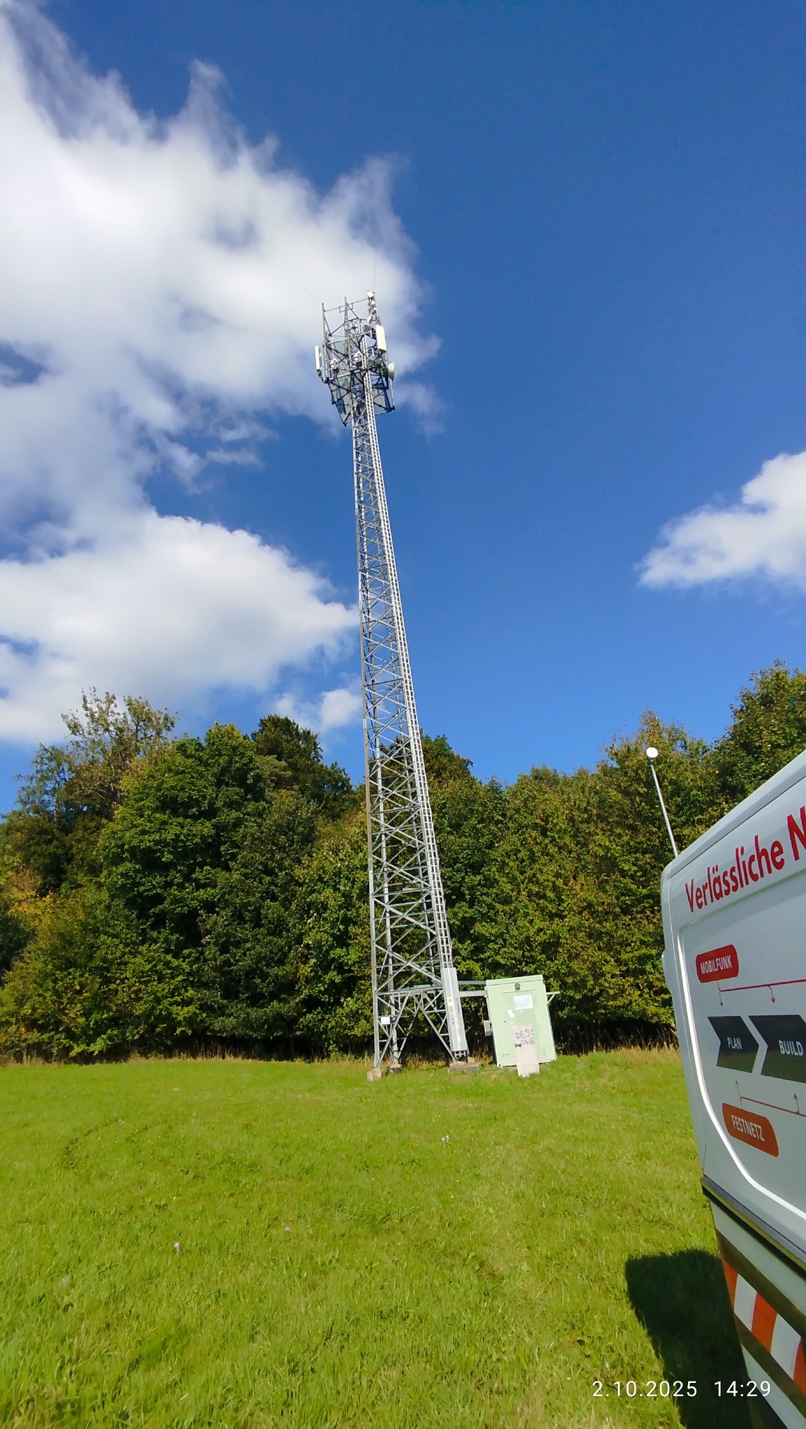 Wysoka wieża telekomunikacyjna z antenami na tle błękitnego nieba z chmurami, widziana z dołu. Obok zaparkowany biały samochód z napisem. Zielona trawa i drzewa w tle.