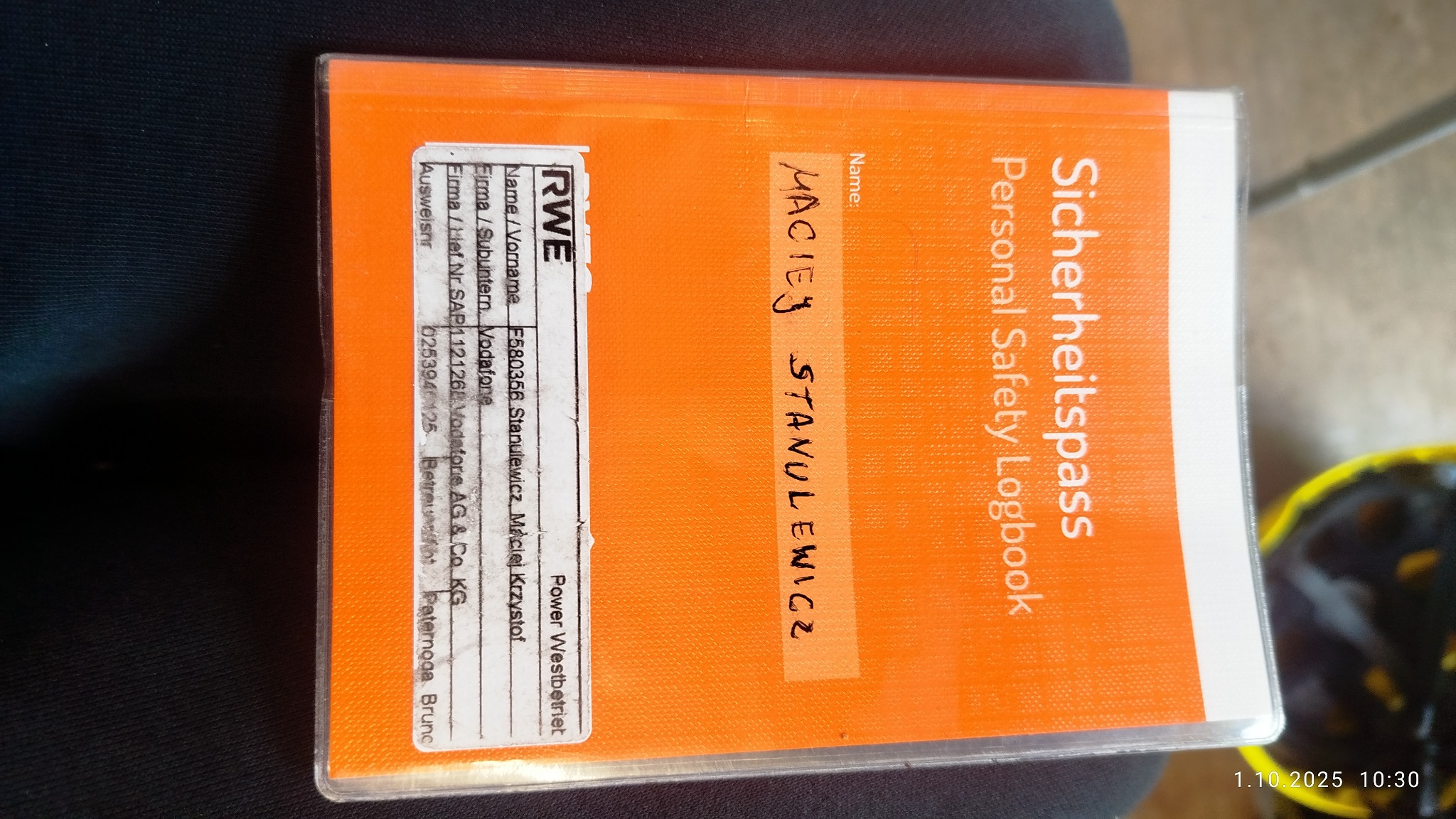 Pomarańczowa książeczka 'Sicherheitspass' z odręcznie wpisanym nazwiskiem 'Maciej Stanulewicz', logo RWE i datą ważności 1.10.2025, leżąca na ciemnym tle.