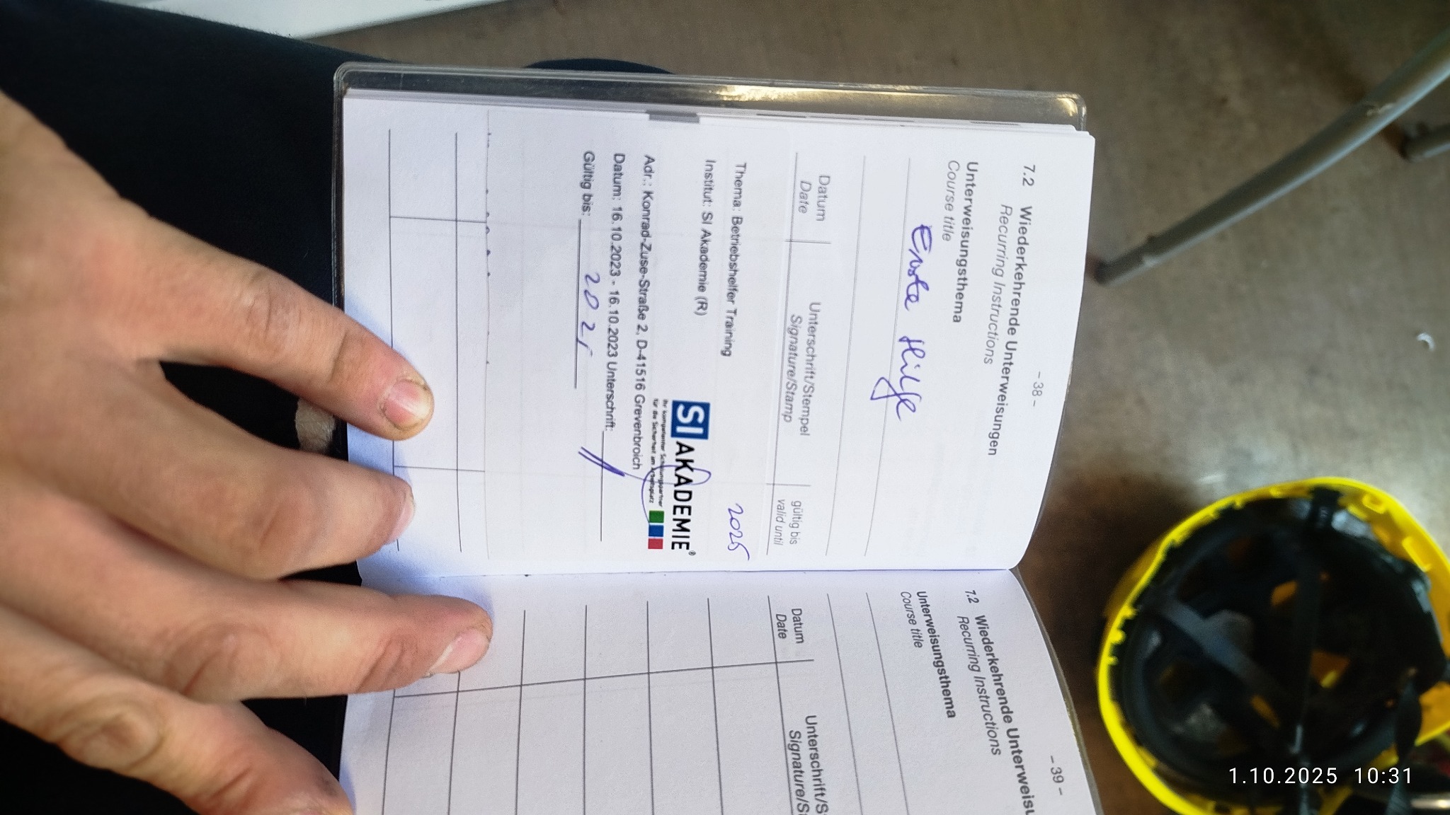 Dokument z podpisem 'Erste Hilfe' i datą ważności 2025, leżący obok żółtego kasku. Widoczne dłonie otwierające dokument. Szkolenie z zakresu bezpieczeństwa.