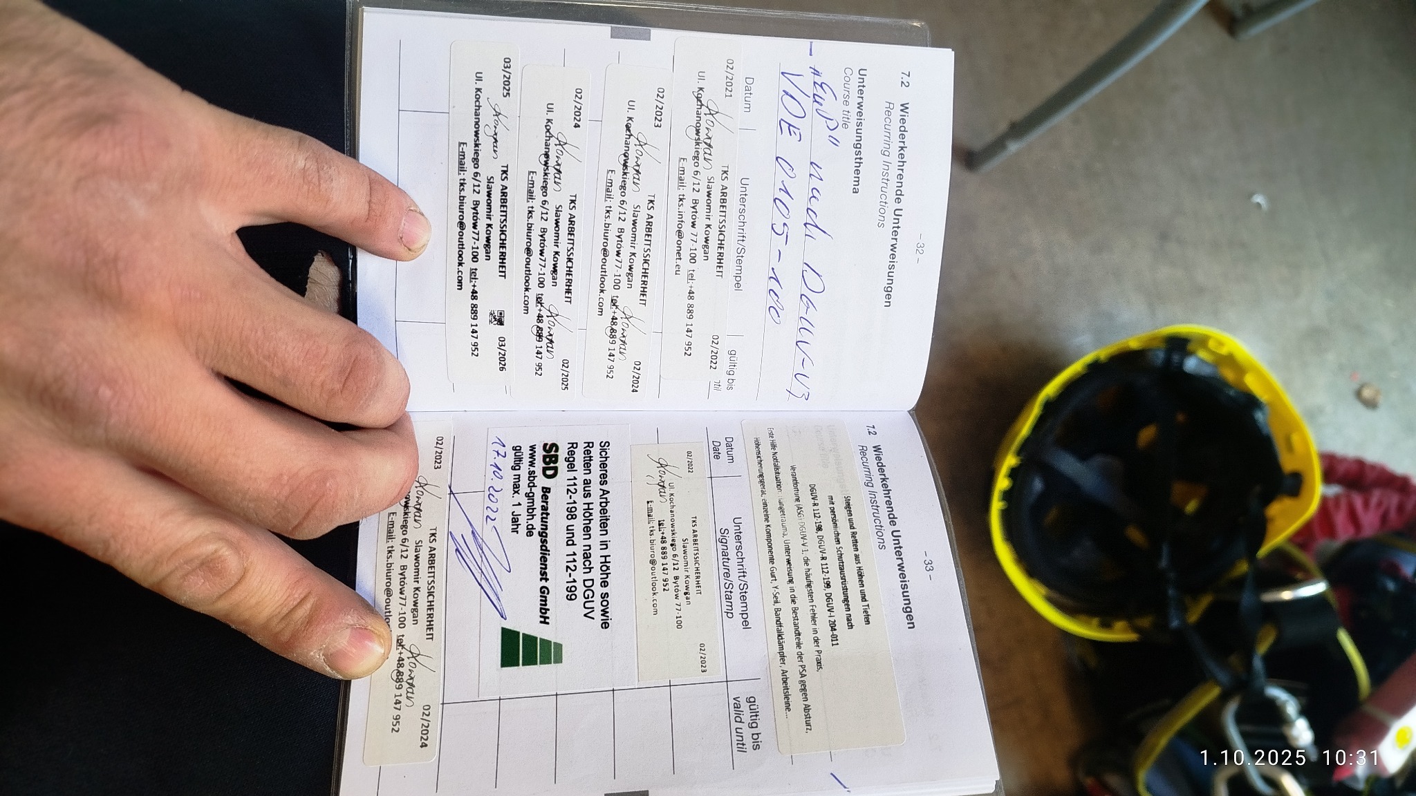 Otwarta książeczka z wpisami szkoleń BHP, widoczne podpisy i pieczątki TKS ARBEITSSICHERHEIT, obok kask ochronny. Dokumentacja potwierdzająca kwalifikacje.