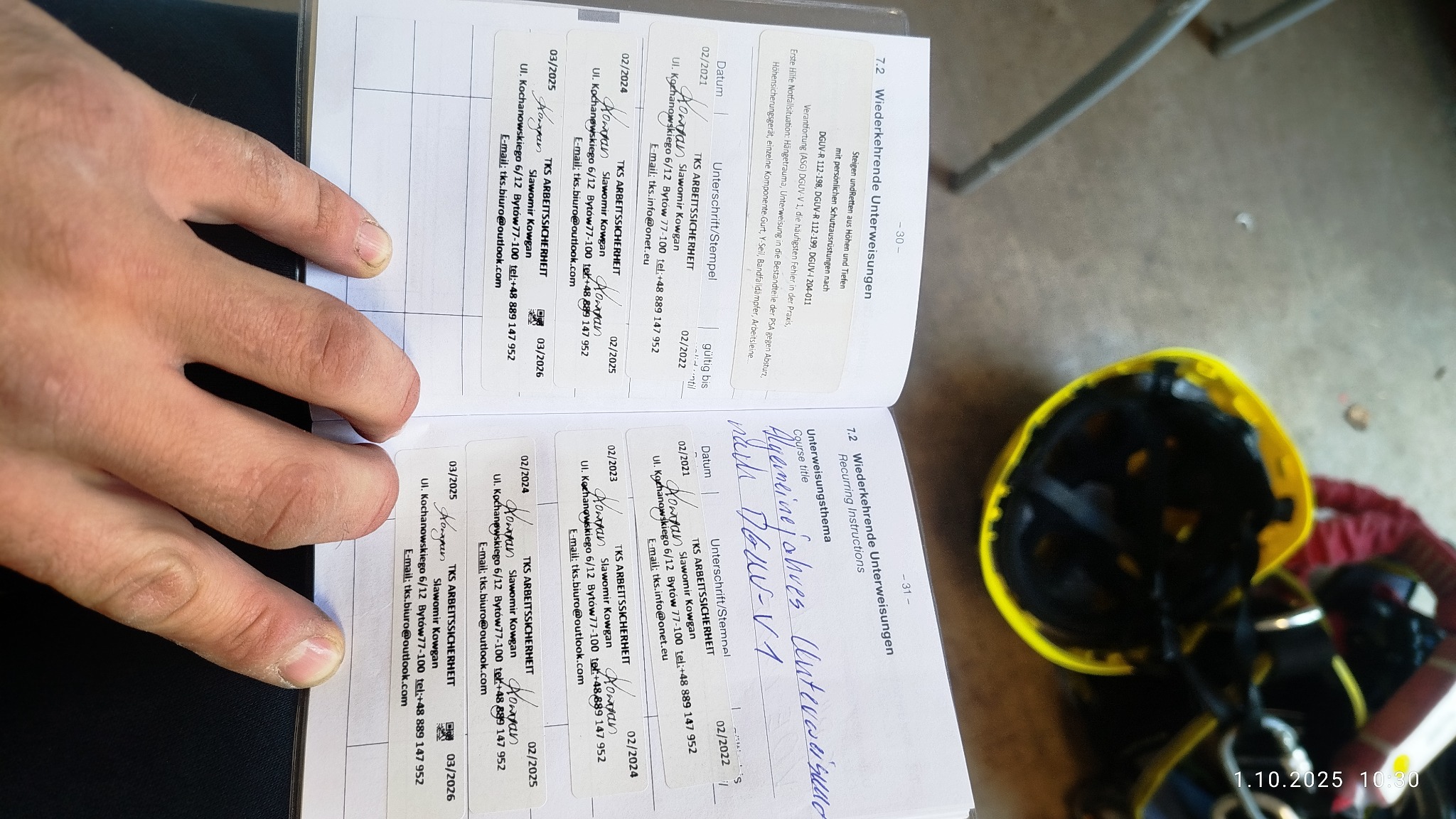 Dokumentacja szkoleń BHP z podpisami, pieczątkami i datami ważności, leżąca obok kasku ochronnego. Widoczne informacje o firmie TKS ARBEITSSICHERHEIT.