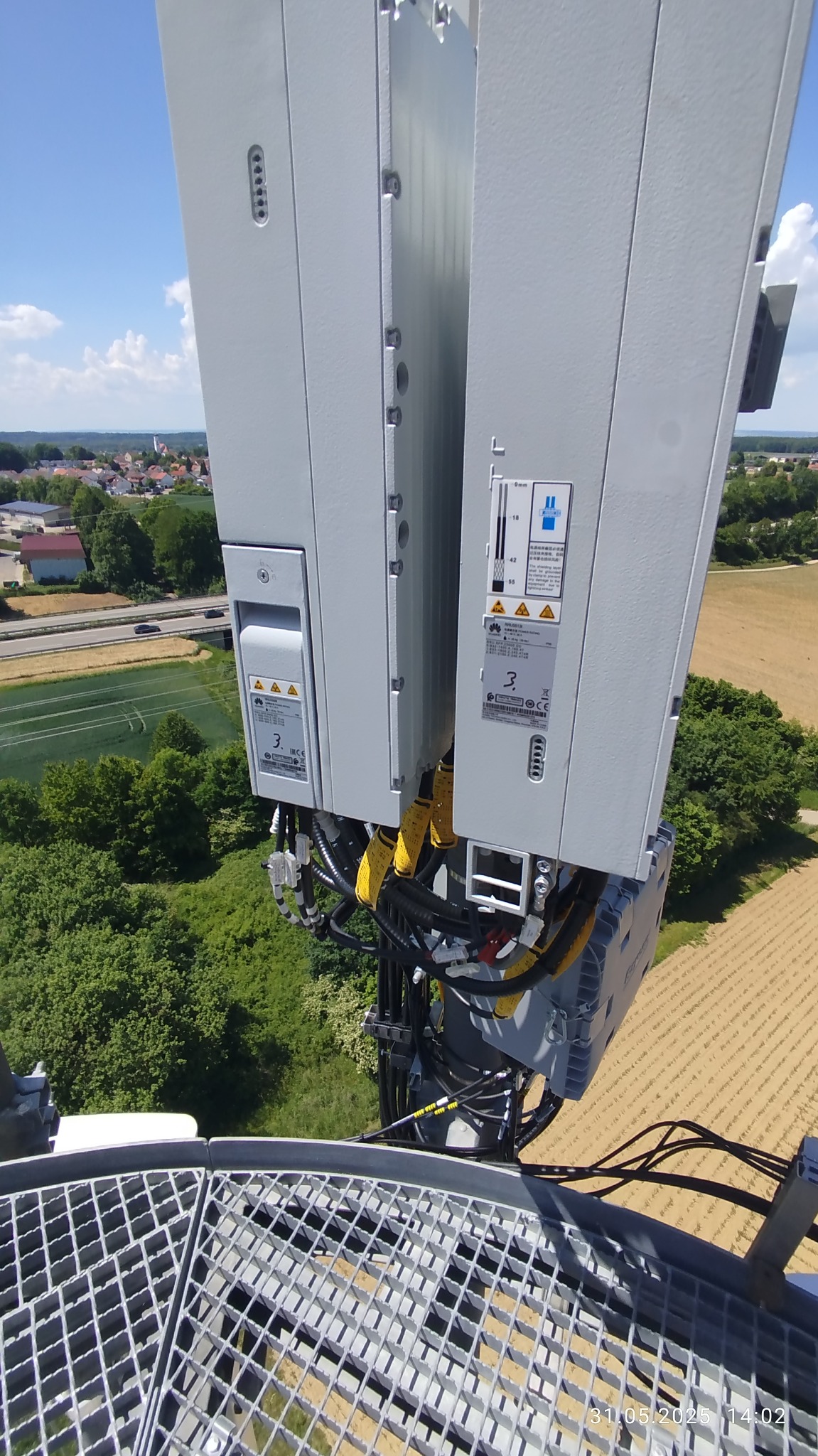 Antena telekomunikacyjna z widocznymi kablami i elementami montażowymi na tle krajobrazu z polami i zabudowaniami. Ujęcie z góry, podkreślające złożoność instalacji.