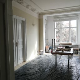 LOS-BUD Łukasz Łosoś - Pokój przed odświeżeniem: zabezpieczona folią podłoga, stół z narzędziami malarskimi, dekoracyjny sufit i okno z widokiem na drzewa. Przygotowania do malowania.