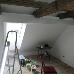 LOS-BUD Łukasz Łosoś - Poddasze po malowaniu na biało, widoczne drewniane belki stropowe, okno dachowe, drabina malarska z lampą, narzędzia malarskie i stolik z czajnikiem.