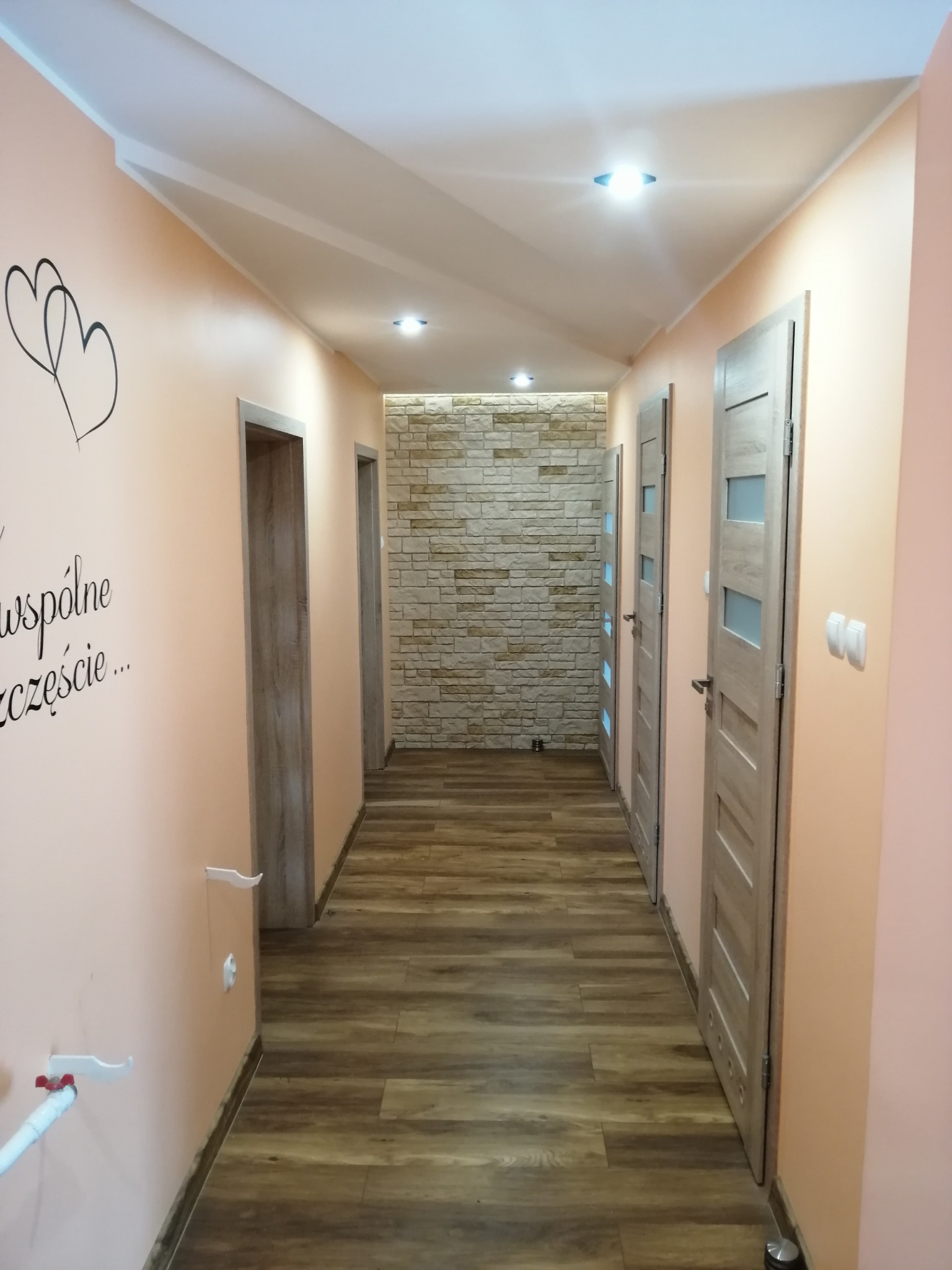 Korytarz po remoncie: ściany w odcieniach brzoskwini z dekoracyjnym napisem, drewnopodobna podłoga, drzwi z przeszkleniami i ściana z kamienia dekoracyjnego.
