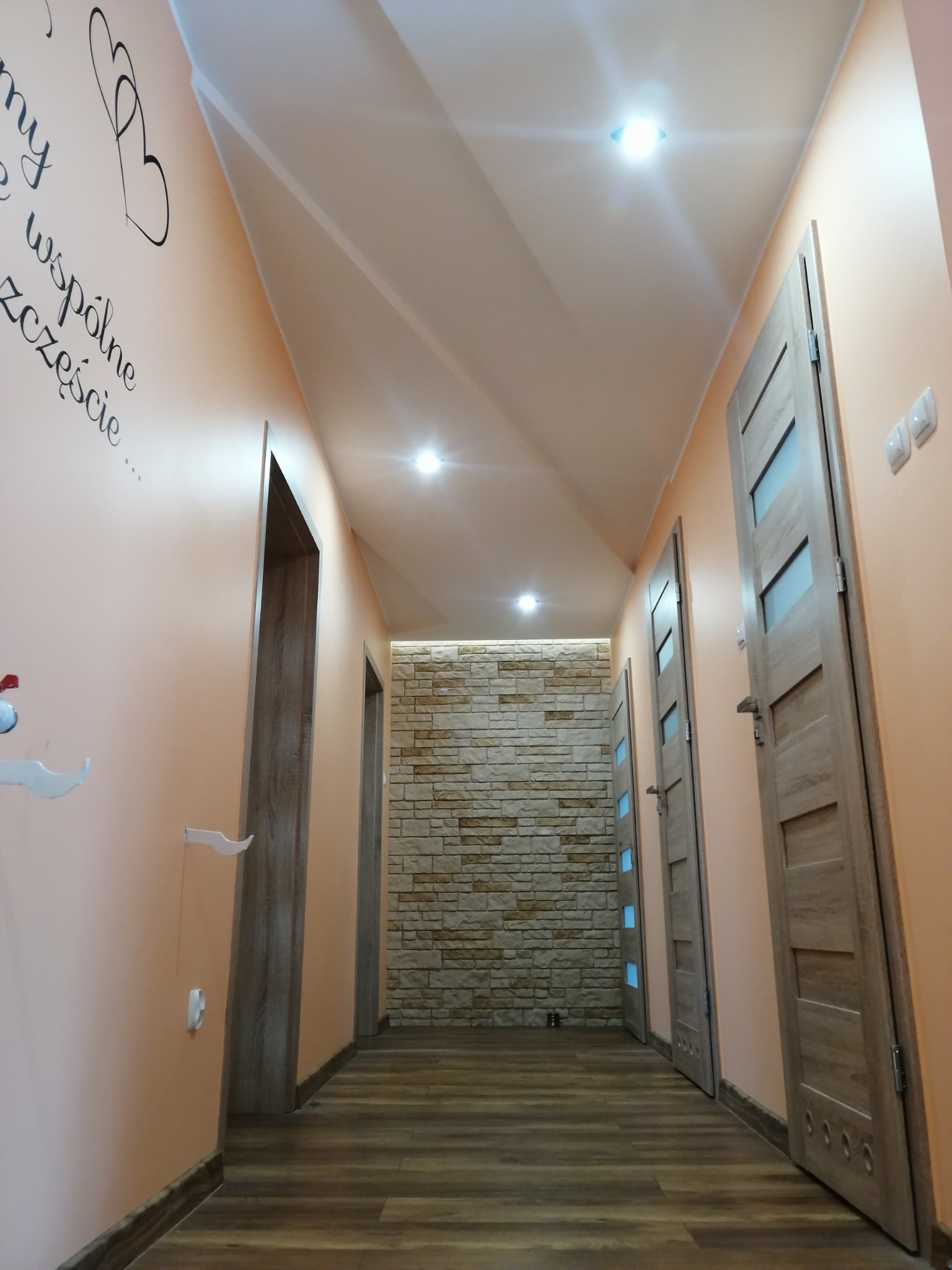 Korytarz po remoncie: ściany w odcieniach brzoskwini z dekoracyjnym napisem, drewnopodobna podłoga, drzwi z przeszkleniami i ściana z kamienia dekoracyjnego w tle.