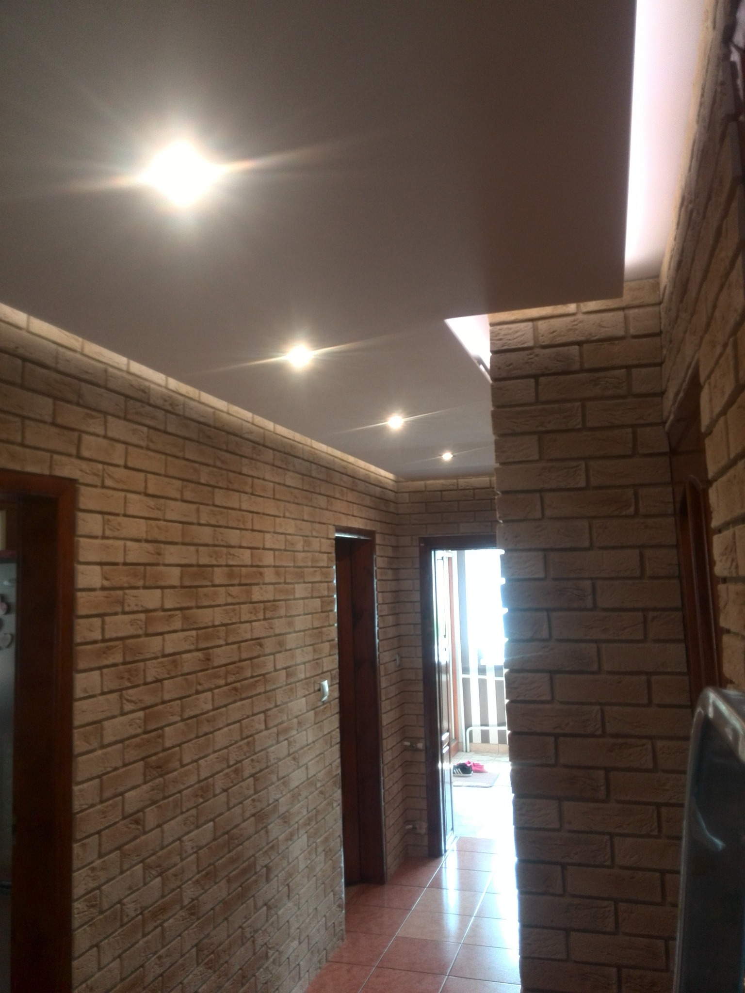 Korytarz z ceglaną ścianą, oświetleniem punktowym w suficie podwieszanym i widocznymi drzwiami. Wykończenie w ciepłych, stonowanych barwach.