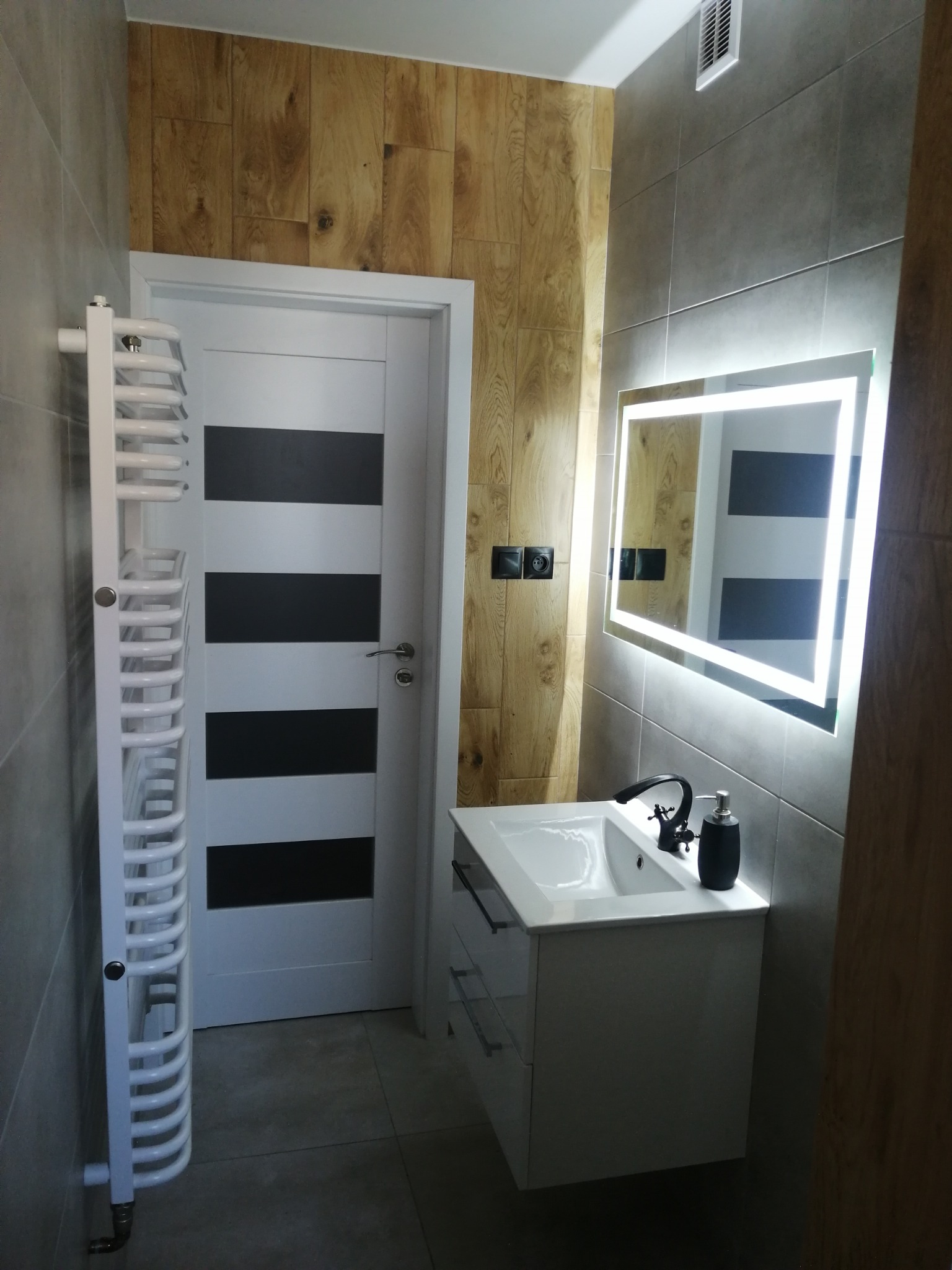 Nowoczesna łazienka po remoncie: szare płytki, drewnopodobna ściana, biała szafka z umywalką, podświetlane lustro i grzejnik drabinkowy. Kontrastowe, czarno-białe drzwi.