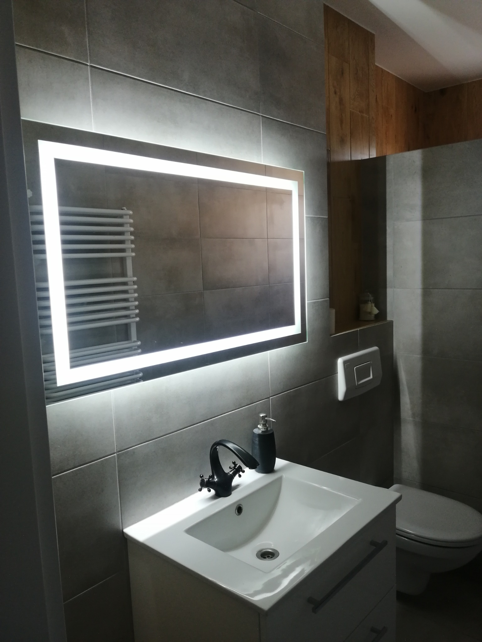 Nowoczesna łazienka z szarymi płytkami, podświetlanym lustrem i białą umywalką z czarną armaturą. Widoczny grzejnik drabinkowy i drewniane akcenty.