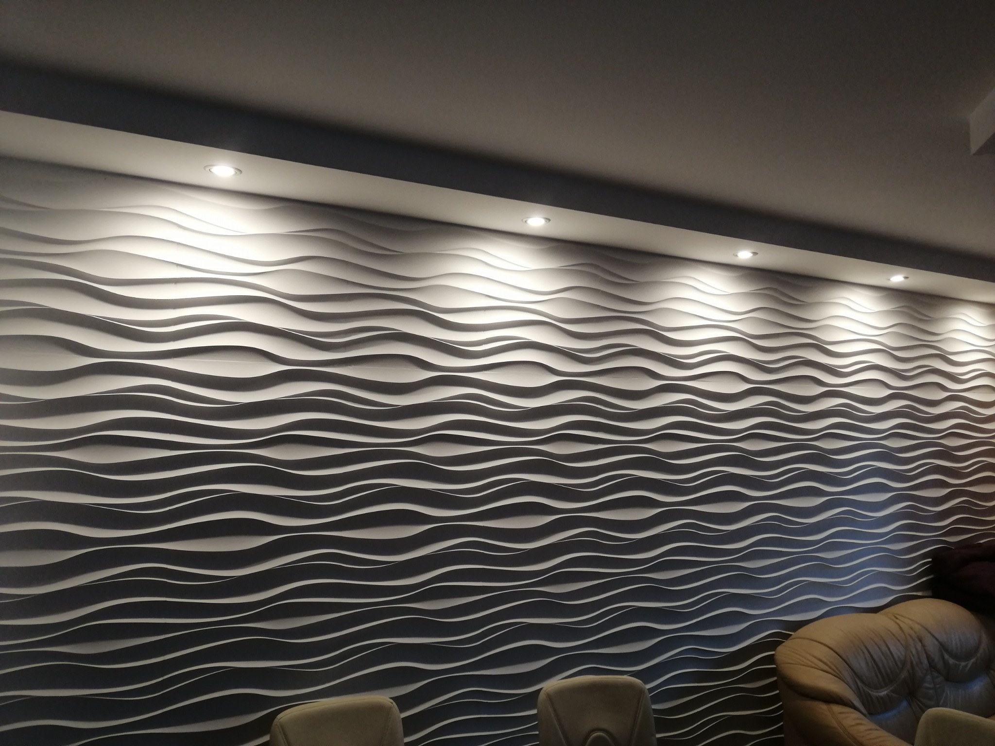 Falista ściana 3D z oświetleniem punktowym, tworząca nowoczesny efekt wizualny. Fragment sofy i krzeseł w dolnej części kadru.