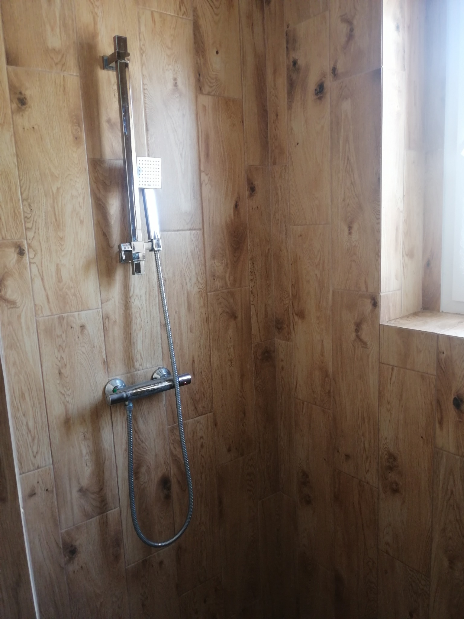 Nowoczesna kabina prysznicowa z drewnopodobnymi płytkami, chromowana armatura, okno z parapetem. Wykończenie łazienki w ciepłych barwach.