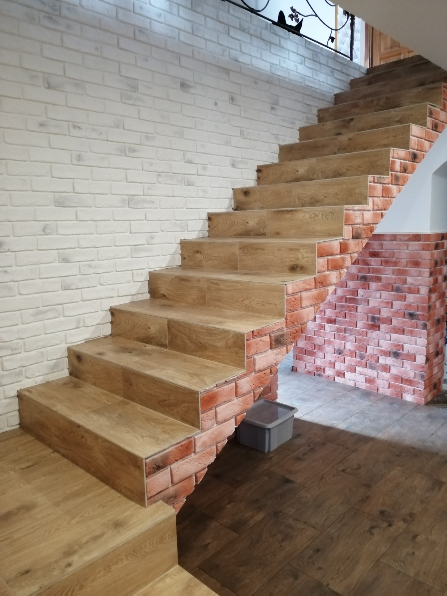 Schody z drewnopodobnymi panelami i ceglaną okładziną, biała ściana z cegły. Widok z dołu, podkreślający detale wykończenia i połączenie materiałów.