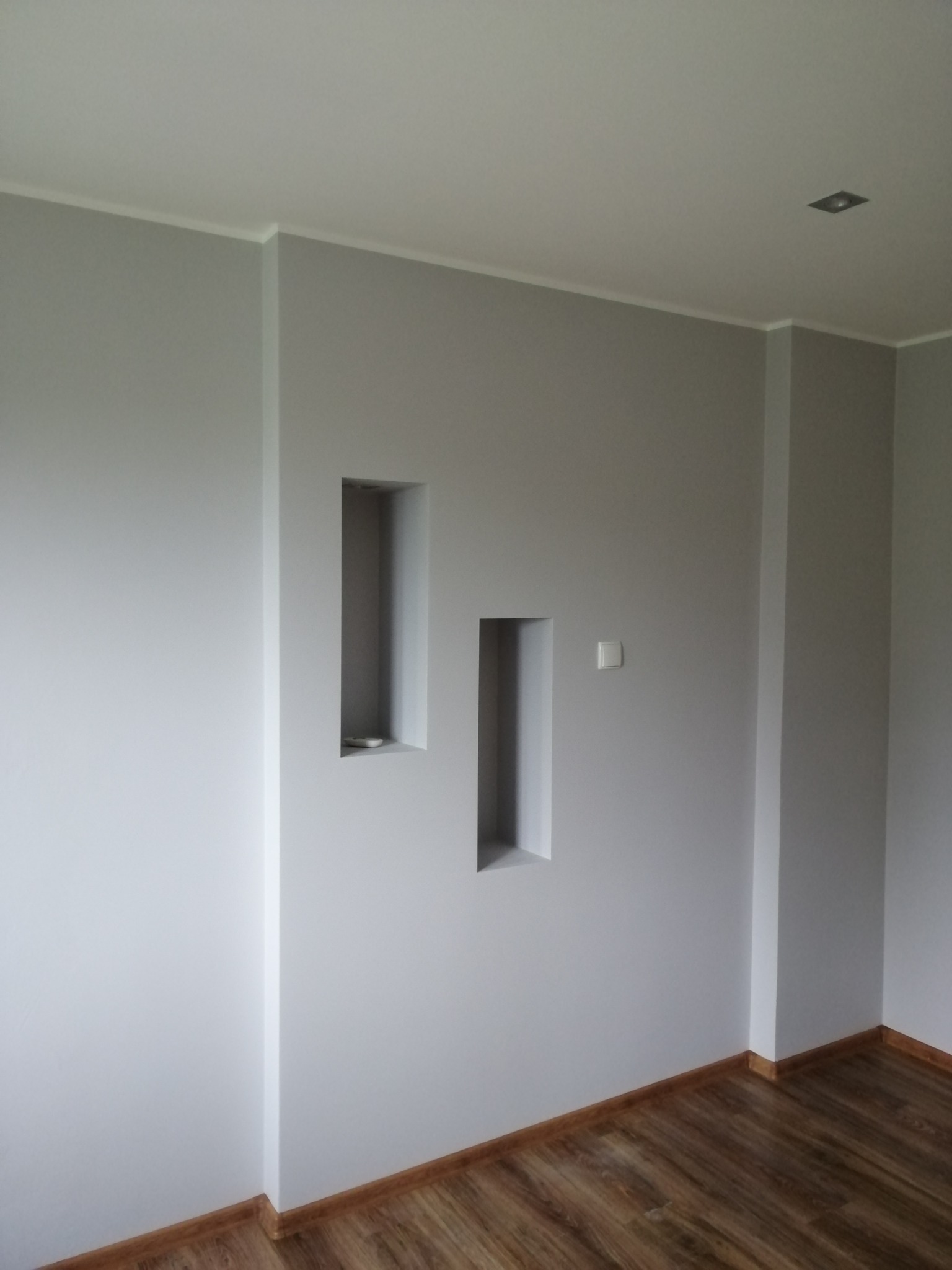 Szare ściany z wnękami i drewnianą podłogą. W jednej wnęce dekoracja. Minimalistyczny design wnętrza z listwami przypodłogowymi i włącznikiem światła.