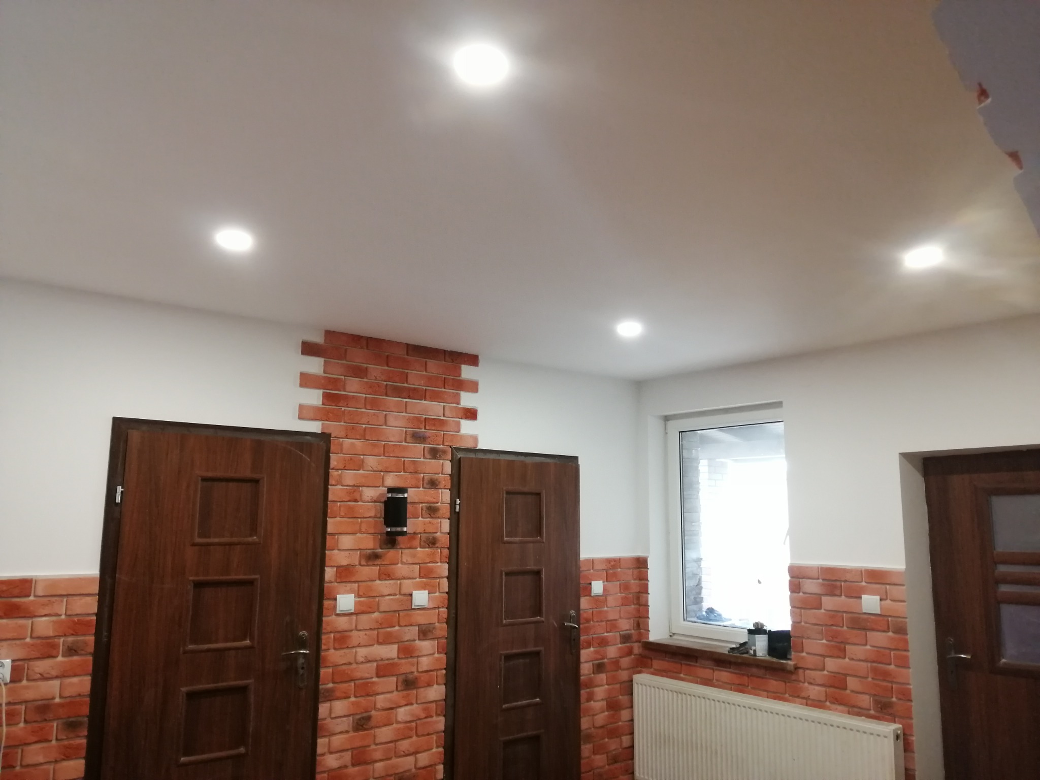 Wykończone wnętrze: ściana z cegły dekoracyjnej, dwie drewniane drzwi, okno z parapetem. Oświetlenie sufitowe punktowe. Grzejnik pod oknem.