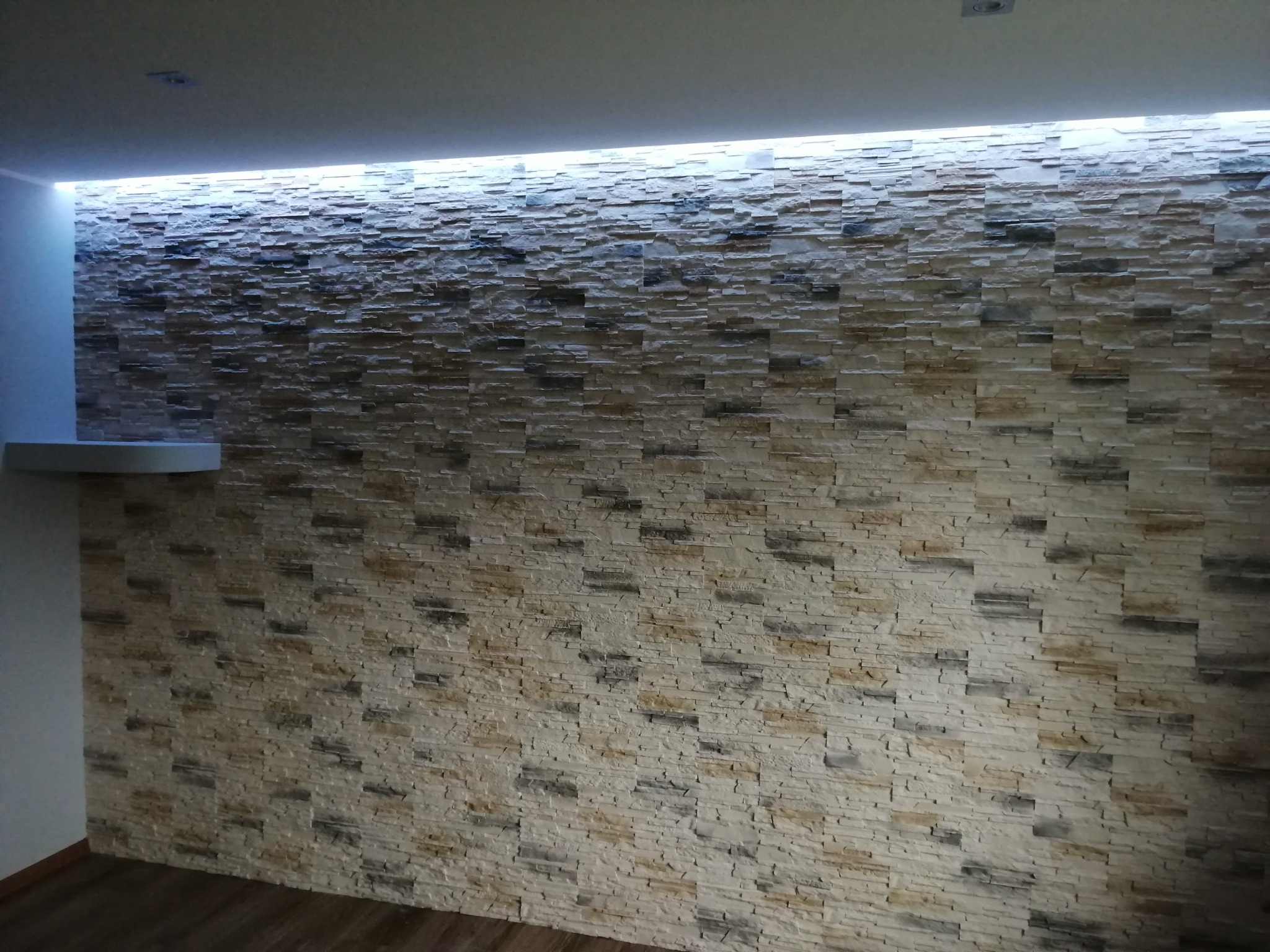 Ściana wyłożona dekoracyjną, beżowo-szarą okładziną kamienną z podświetleniem LED u góry, widoczna z perspektywy pod lekkim kątem. Fragment podłogi z paneli.