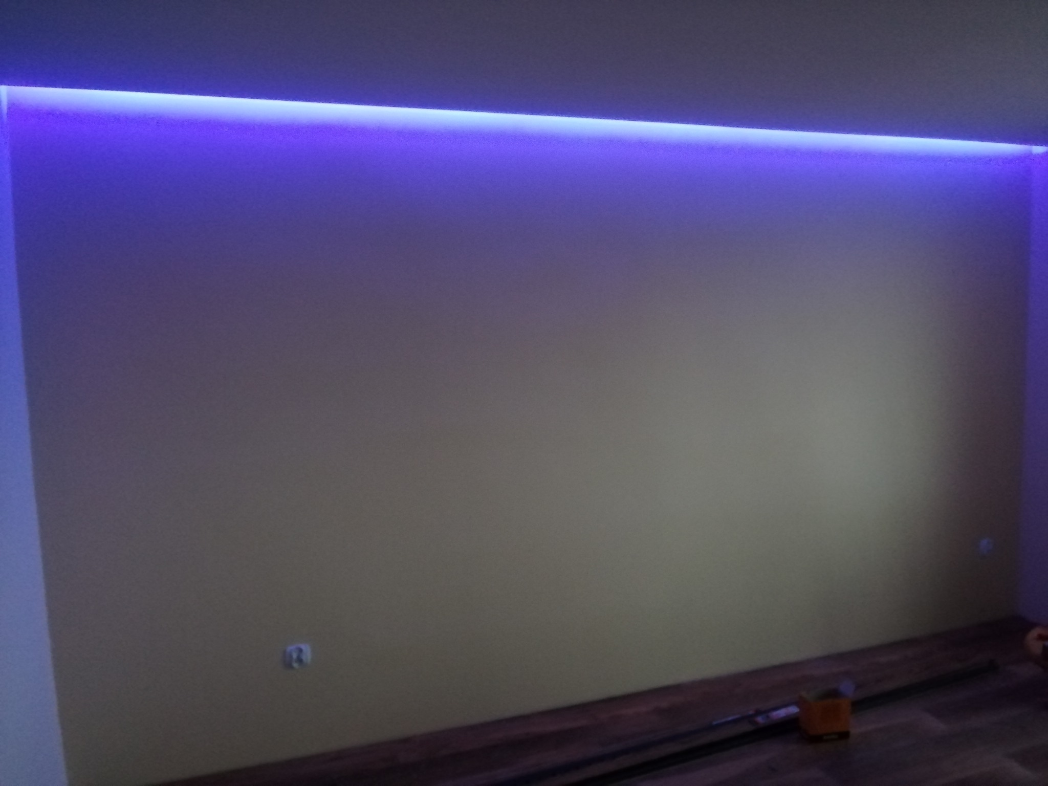 Pokój z podświetleniem LED w suficie, ściany w odcieniach beżu, panele podłogowe. Widoczny fragment gniazdka elektrycznego i narzędzia na podłodze.