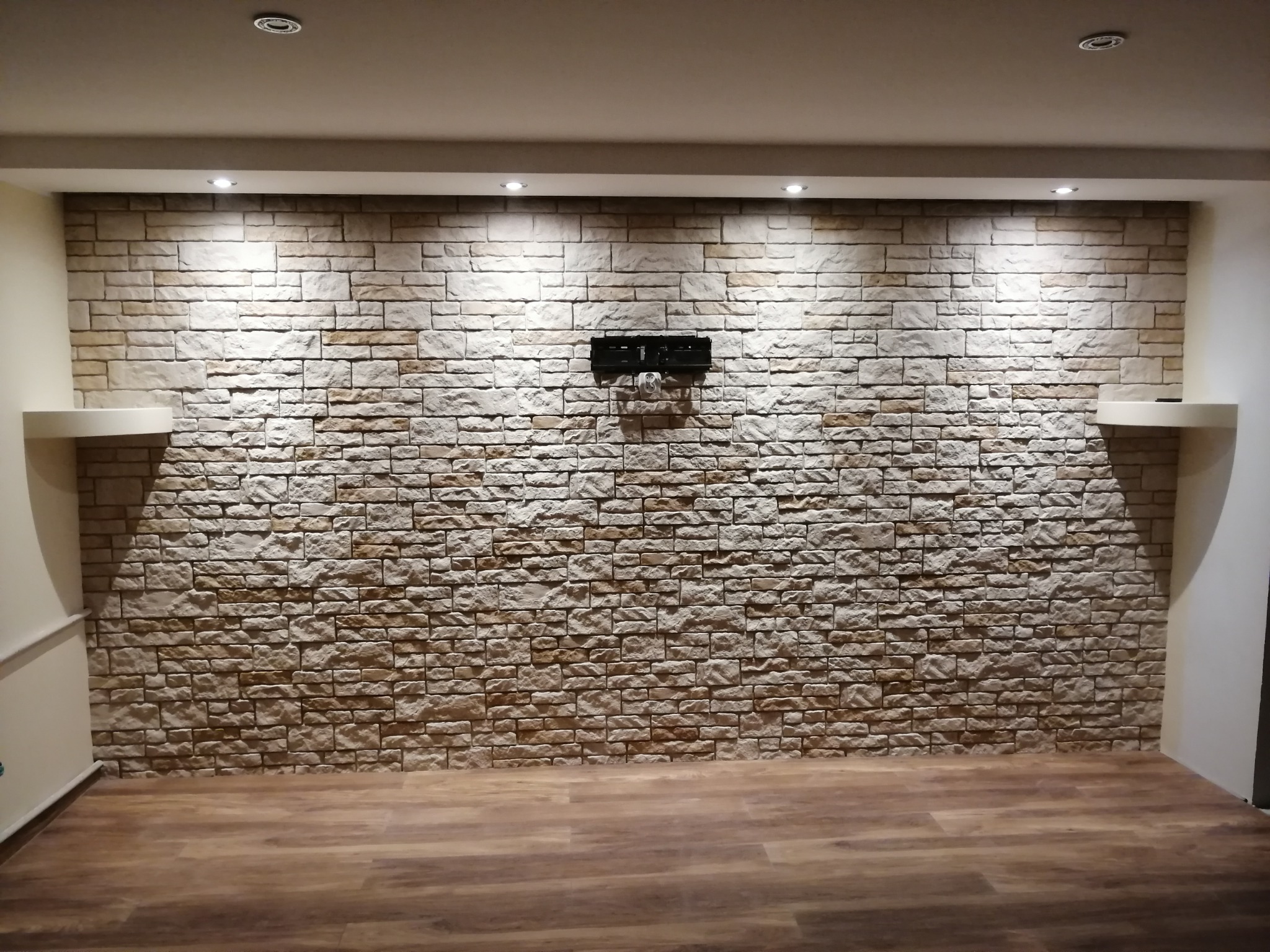 Ściana z dekoracyjnej cegły z uchwytem na telewizor i półkami, oświetlona punktowo, na podłodze panele. Wykończenie wnętrza w ciepłych barwach.