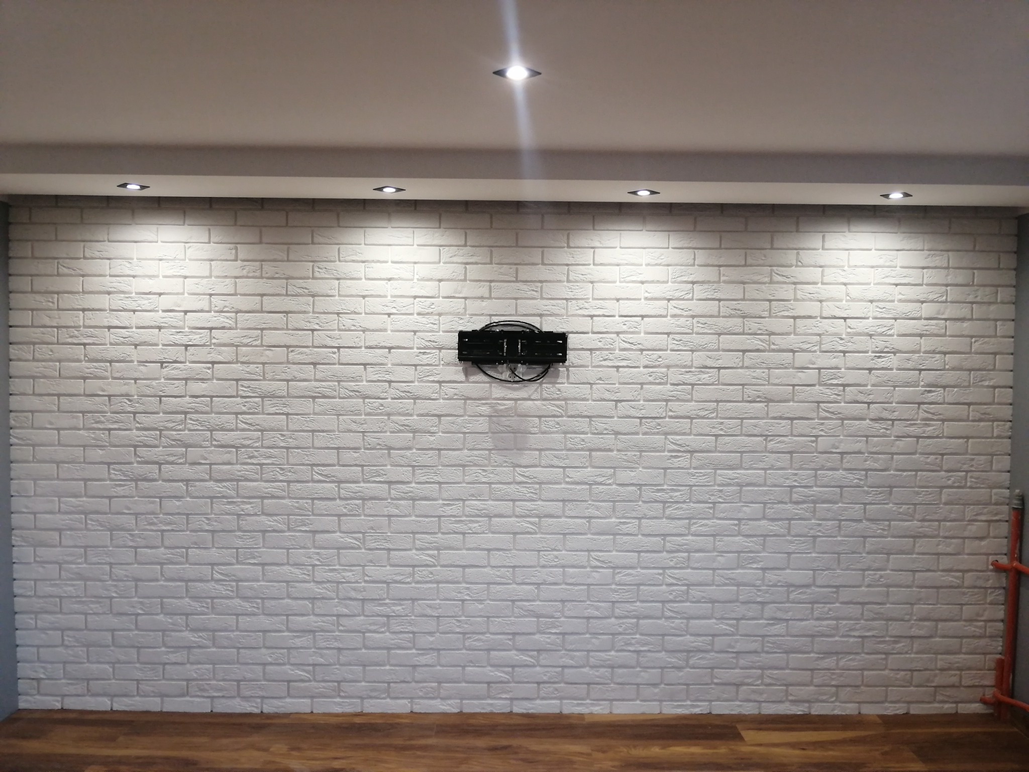 Biała ściana z cegły dekoracyjnej, oświetlona punktowo, z zamontowanym uchwytem na telewizor. Widoczna drewniana podłoga i fragment instalacji grzewczej.