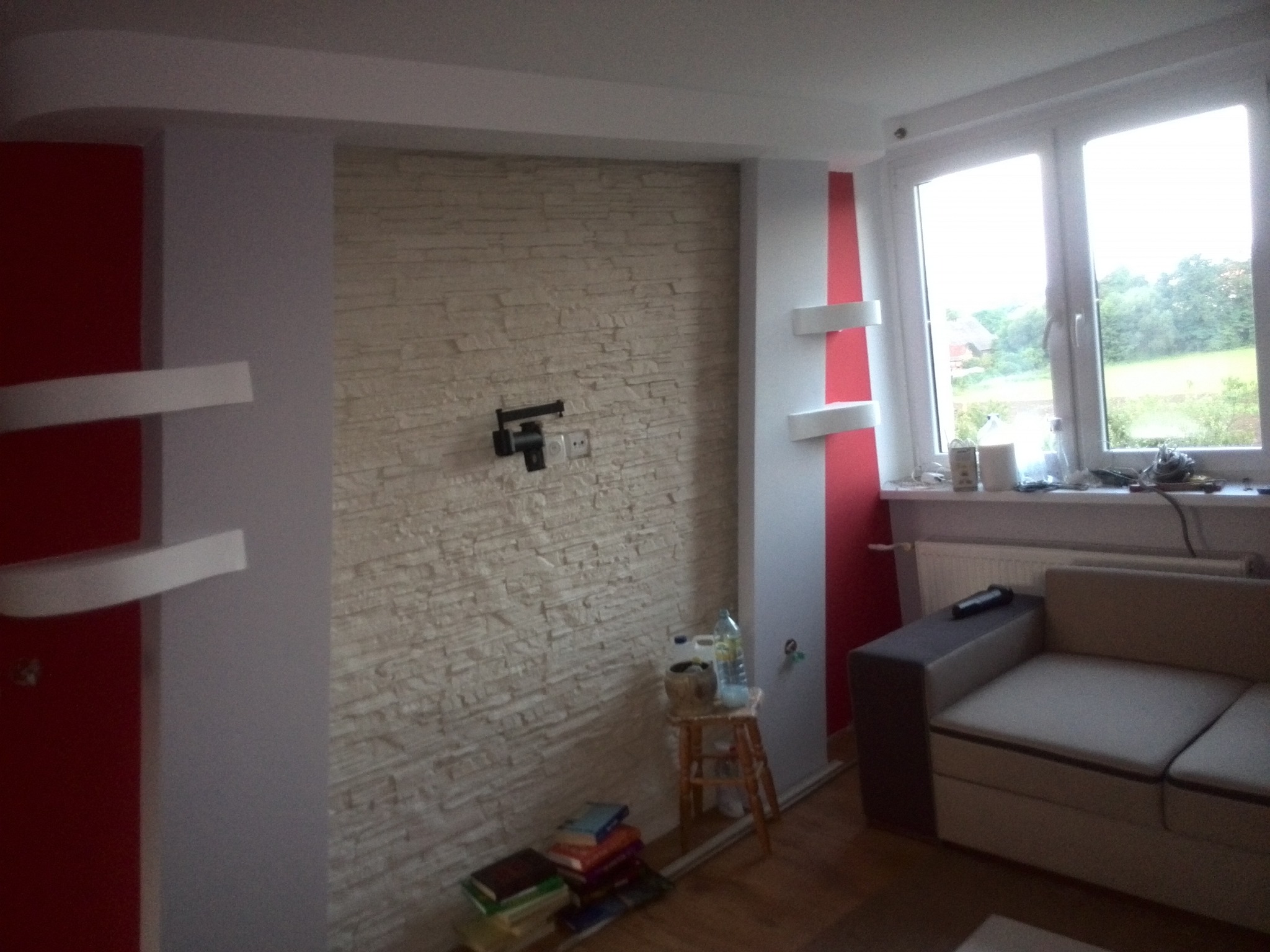 Jasne wnętrze pokoju z dekoracyjną ścianą z kamienia, nowoczesnymi półkami i dużym oknem. Widoczny fragment sofy i stolik z książkami. Ściana w odcieniach beżu.