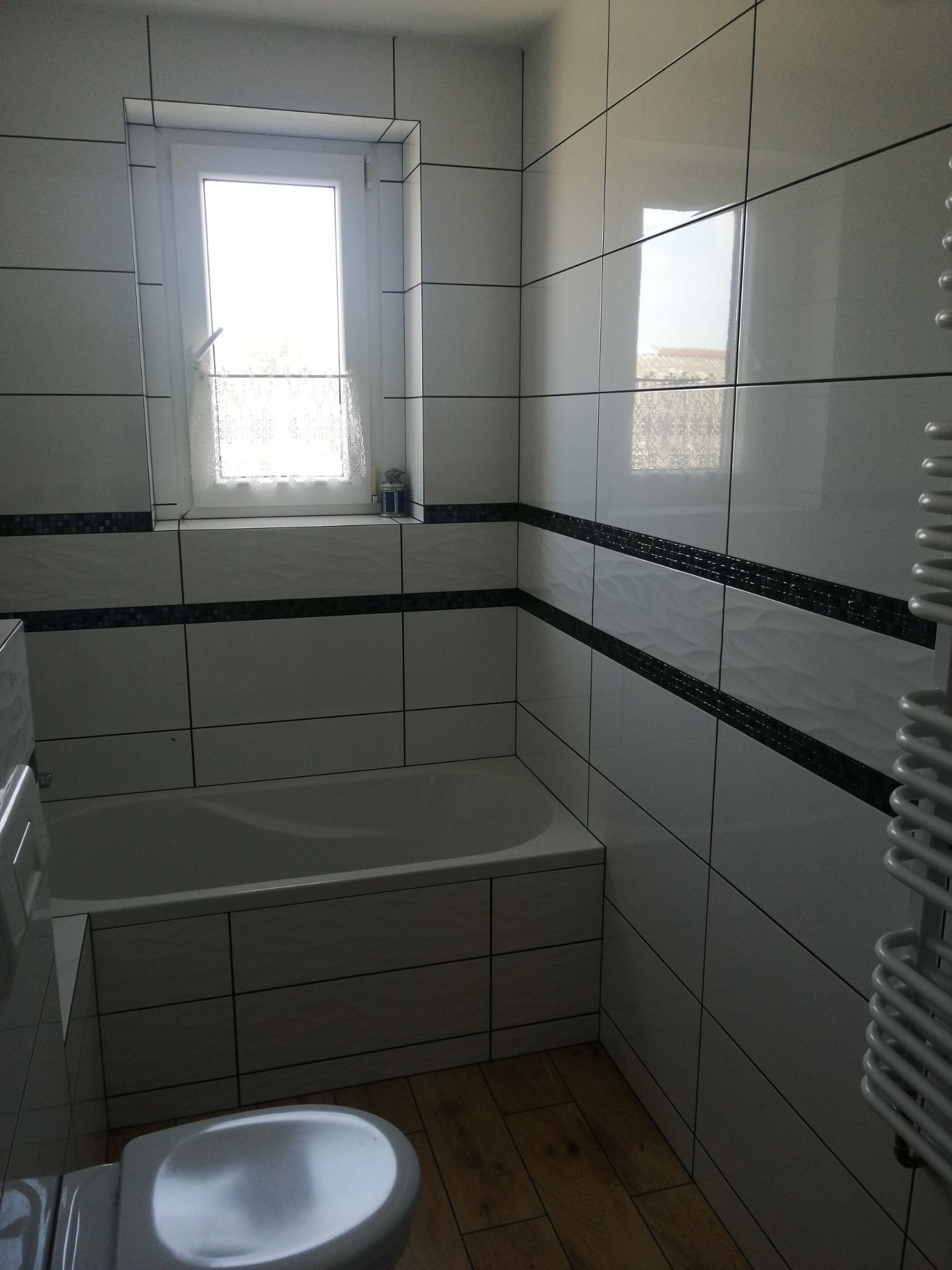 Wykończona łazienka z biało-czarnymi płytkami, wanną obudowaną płytkami i grzejnikiem drabinkowym. Widoczne okno z firanką i drewnopodobna podłoga.