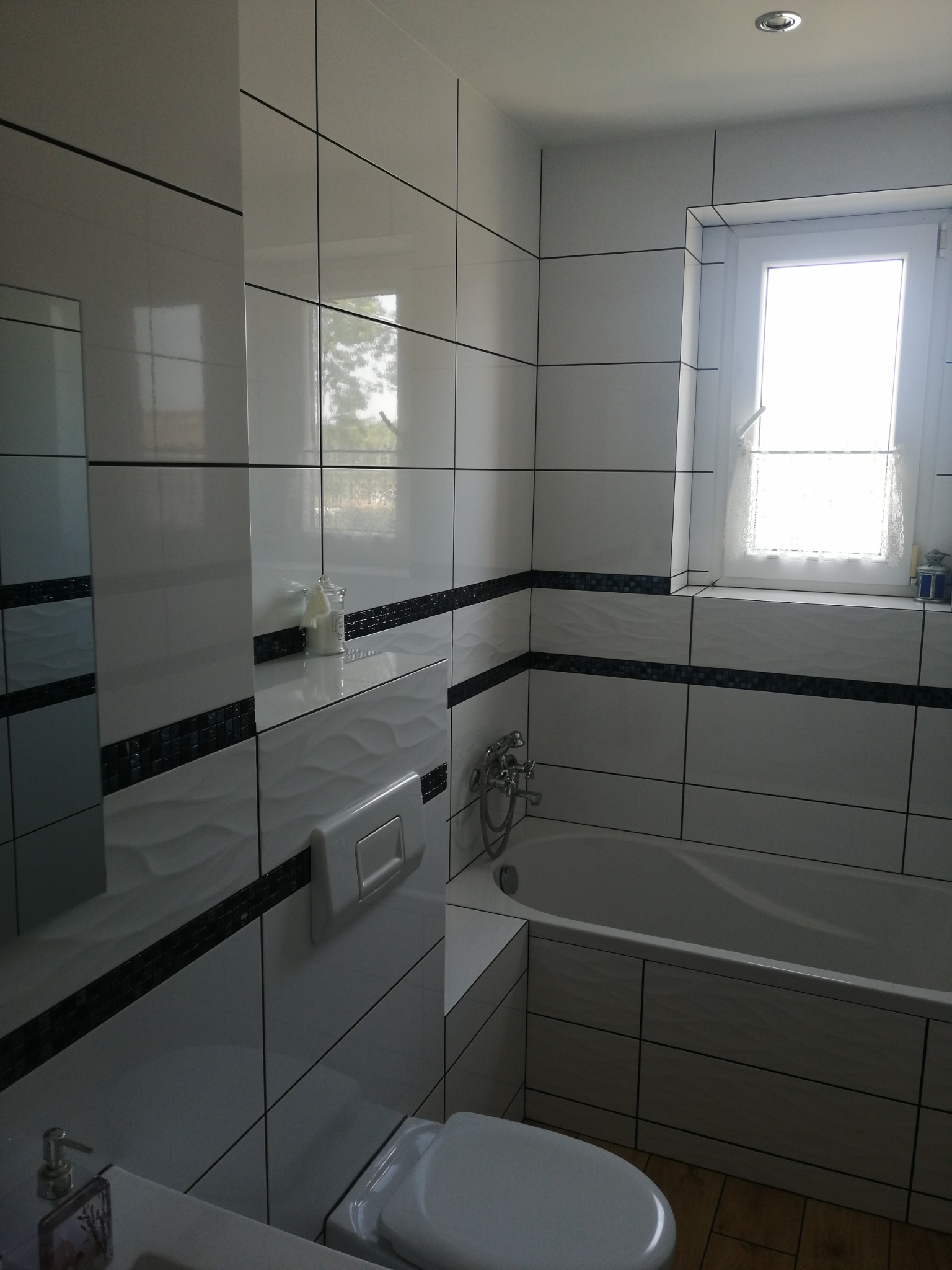 Biała łazienka z wanną, wykończona błyszczącymi płytkami z czarnym akcentem. Widoczne okno z firanką i detale armatury. Umywalka z dozownikiem w rogu kadru.