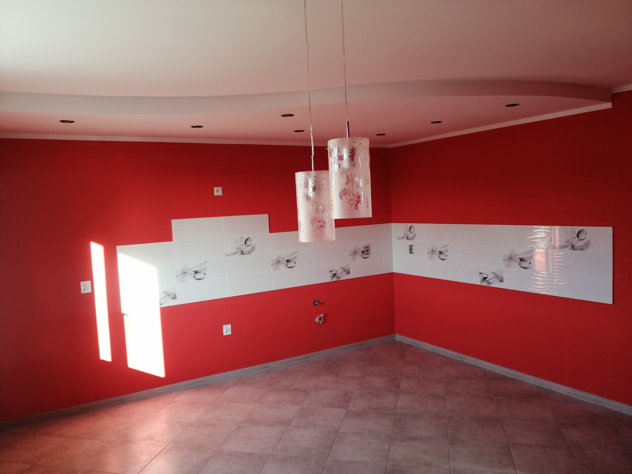 Czerwona kuchnia po remoncie: białe płytki z motywem kwiatowym nad blatem, nowoczesne lampy wiszące, gładkie ściany i podłoga z płytek. Jasne, przestronne wnętrze.