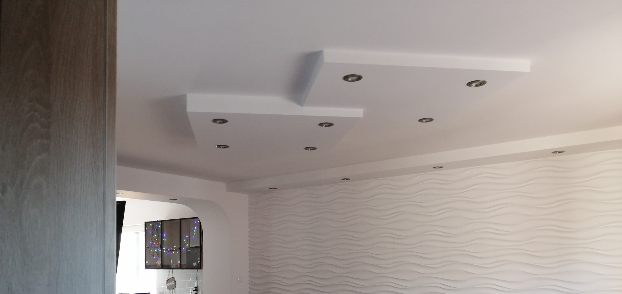 Biały sufit podwieszany z oświetleniem punktowym, dekoracyjna ściana z panelami 3D w tle. Widok z dołu, fragment szafy po lewej stronie kadru.