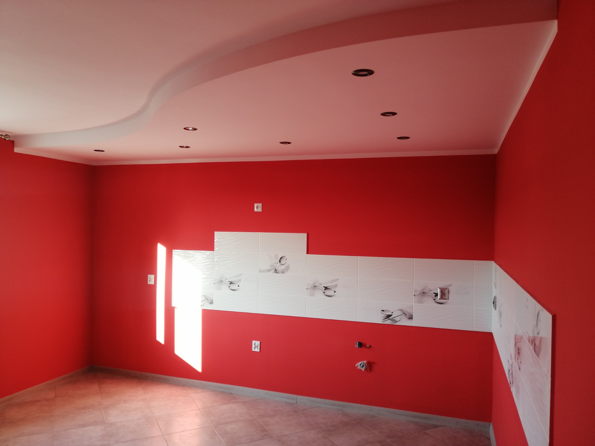 Czerwona kuchnia po remoncie: białe płytki z motywem sztućców, gładkie ściany, podwieszany sufit z oświetleniem punktowym i jasna podłoga z płytek. Widok na fragment pomieszczenia.