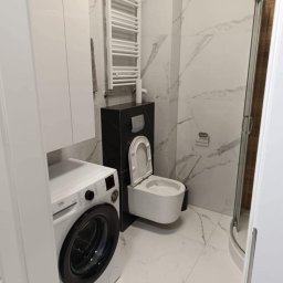 Instalatorstwo Sanitarne Ivan Tkachuk - Nowoczesna łazienka z marmurowymi płytkami, wiszącą toaletą z czarnym stelażem, grzejnikiem drabinkowym i pralką. Minimalistyczny design, jasne wnętrze.
