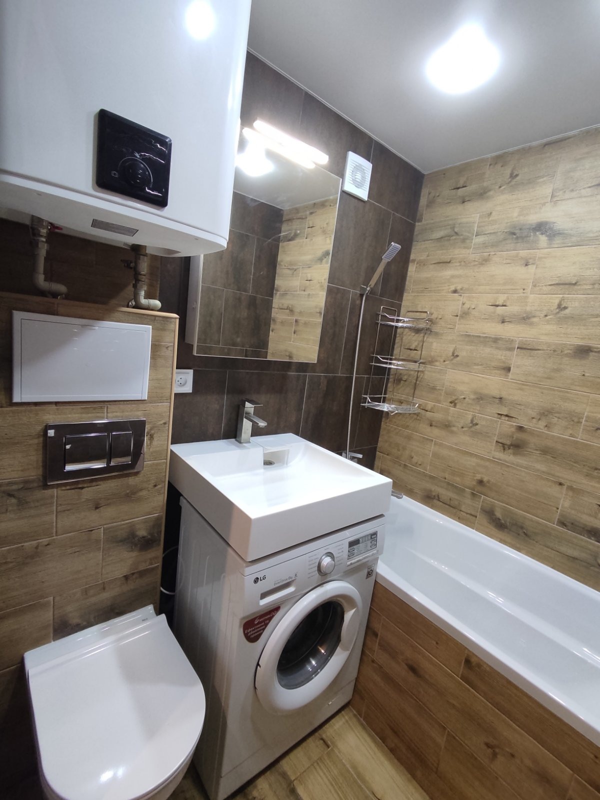 Nowoczesna łazienka z drewnopodobnymi płytkami, białą armaturą i pralką pod umywalką. Widoczny bojler i bateria umywalkowa. Oświetlenie sufitowe.