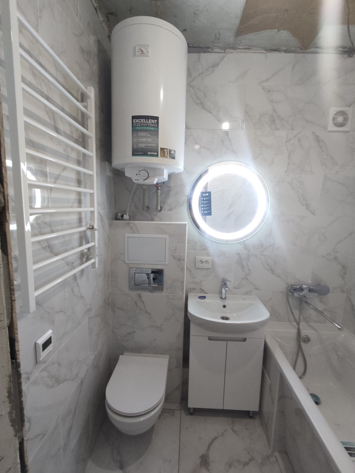 Biała łazienka z marmurowymi płytkami, podgrzewacz wody nad toaletą, okrągłe lustro z podświetleniem LED, umywalka z szafką i grzejnik drabinkowy.