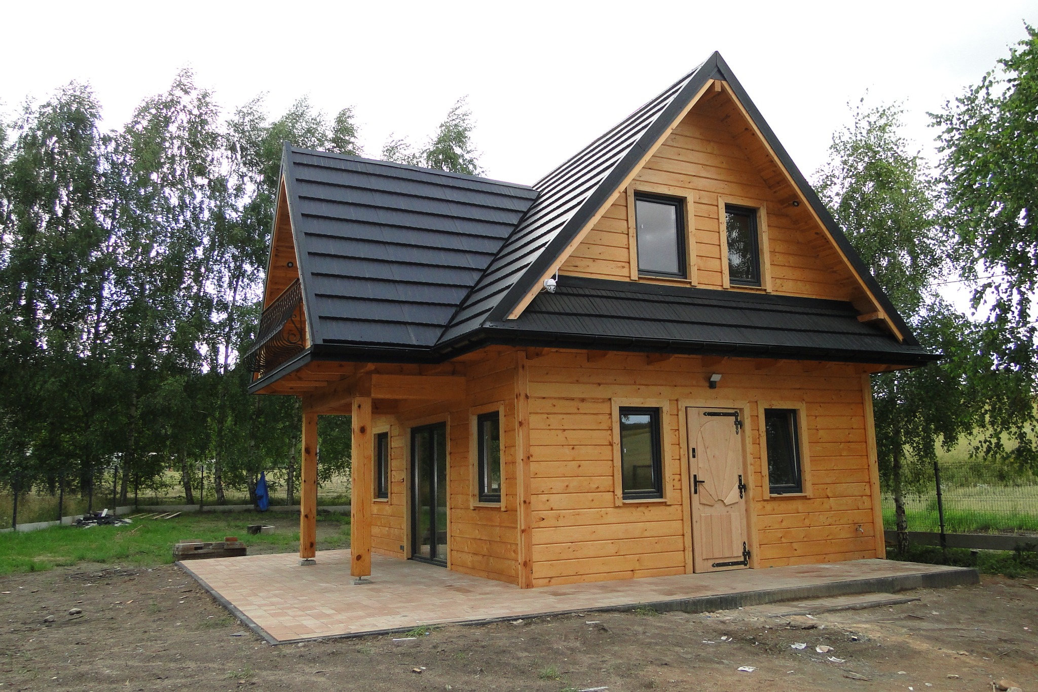 Drewniany domek letniskowy z czarnym dachem, widok z zewnątrz. Jasne drewno, ciemne okna i drzwi. Balkon z ozdobną balustradą. Otoczenie: trawa i drzewa.