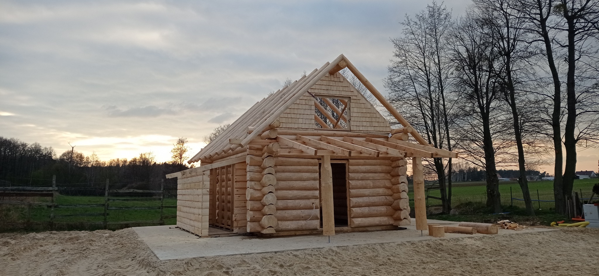 Surowy stan budowy domu z bali, widoczne drewniane ściany, dach z desek i elementy konstrukcyjne. Tło stanowią drzewa i zachodzące słońce.