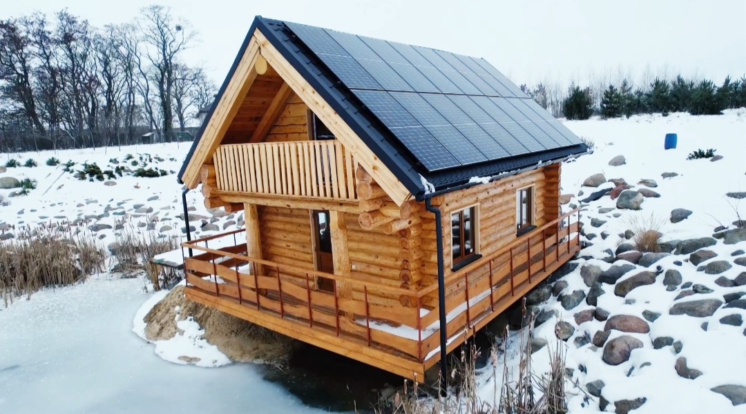 Drewniany domek z bali z panelami słonecznymi na dachu, częściowo zanurzony w zamarzniętym stawie, otoczony śniegiem i kamieniami. Widok z boku, pochmurny dzień.