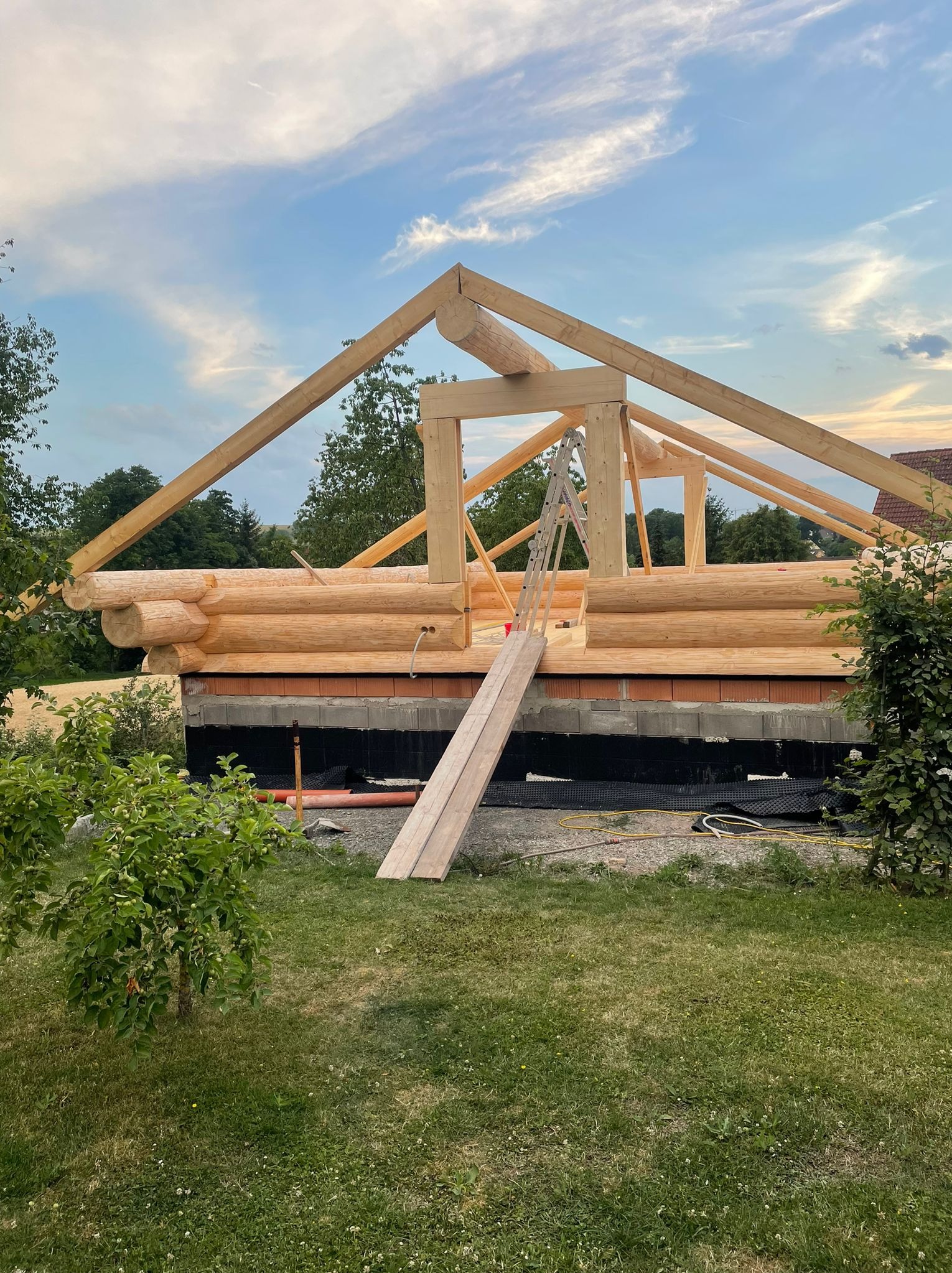 Wznoszenie domu z bali: surowe drewno, widoczne połączenia bali, fragment dachu, drabina oparta o konstrukcję, fundament z cegły i izolacji, trawa, drzewa w tle.