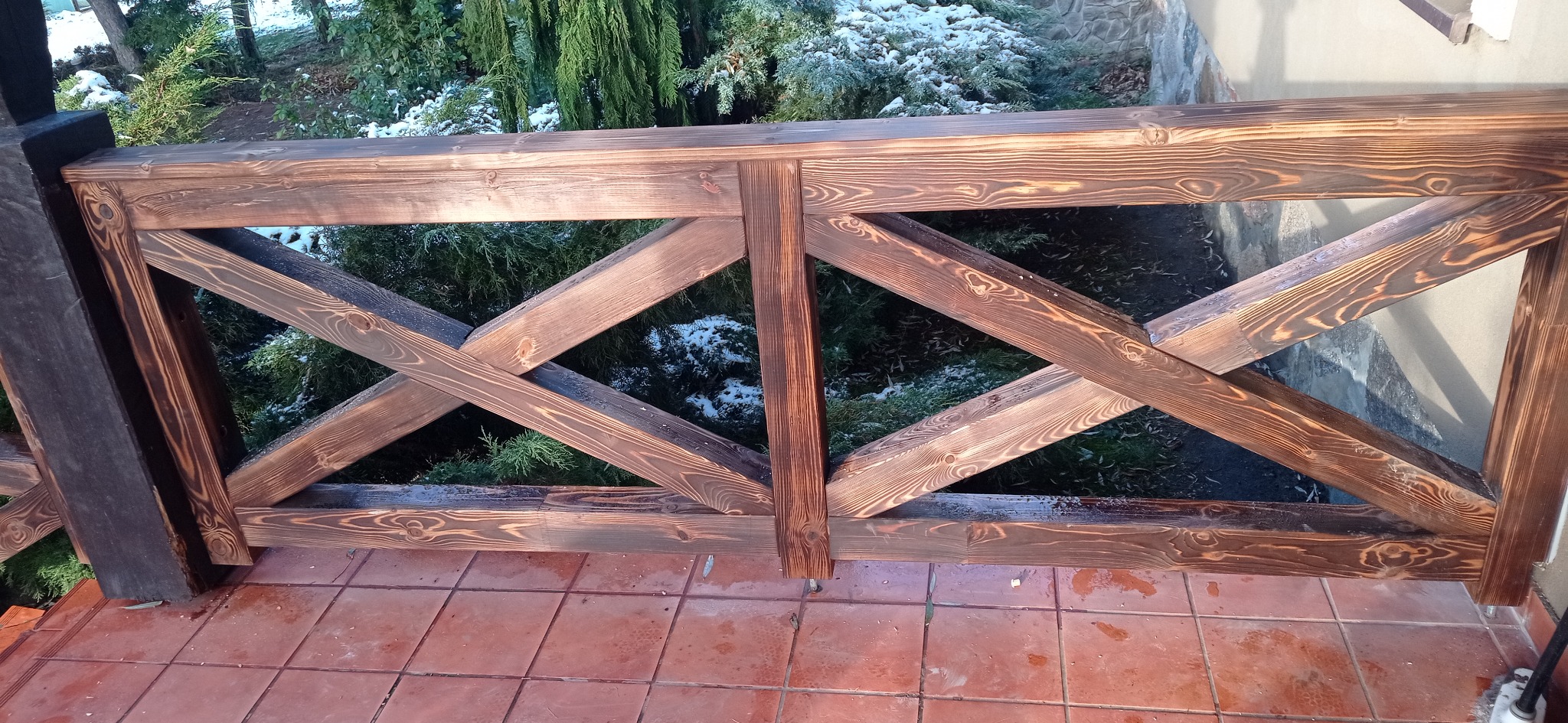 Drewniana balustrada tarasu z krzyżowym wzorem, bejcowana na ciemny brąz, na tle zieleni i częściowo ośnieżonego ogrodu. Widok z bliska, podkreślający strukturę drewna.