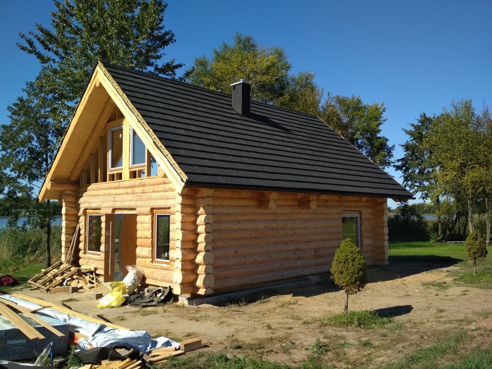 Surowy dom z bali z czarnym dachem na tle zieleni i błękitnego nieba. Widoczne elementy konstrukcyjne i materiały budowlane. Etap budowy, jasne drewno.