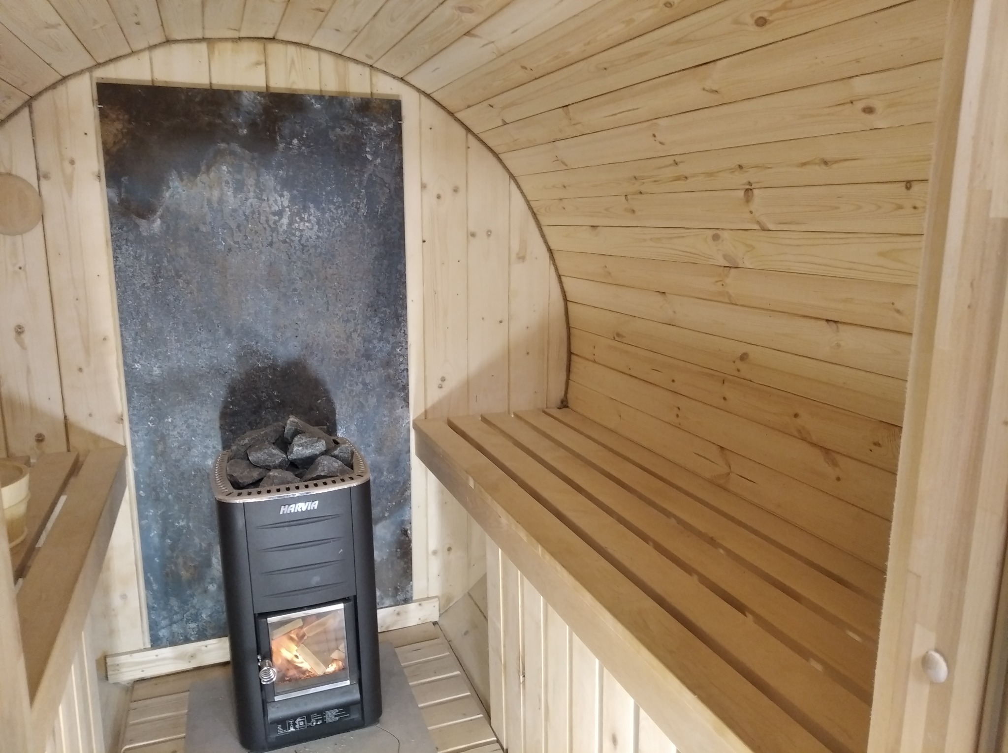Drewniana sauna z piecem Harvia i kamienną ścianą. Jasne drewno, ławy z listew, piec z kamieniami. Przytulne wnętrze sauny.